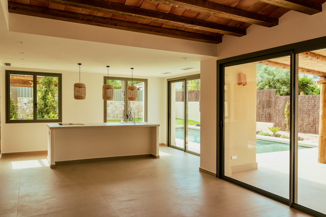 Offener Wohnbereich mit Sichtbalkendecke, minimalistischer Kücheninsel mit Rattanhängeleuchten und großen Glasschiebetüren zum privaten Poolbereich.