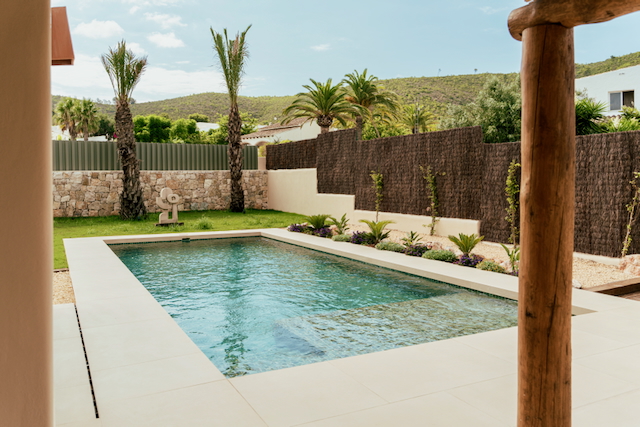 Rechteckiger Pool mit hellen Feinsteinzeugfliesen, umgeben von einem Garten mit Palmen, Natursteinmauern und Blick auf die Berge.