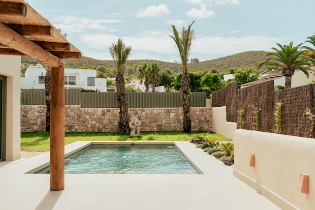 Rechteckiger Swimmingpool auf einer Feinsteinzeug-Terrasse, umgeben von Natursteinmauern, mediterranem Garten mit Palmen und einer rustikalen Holzbalken-Veranda.