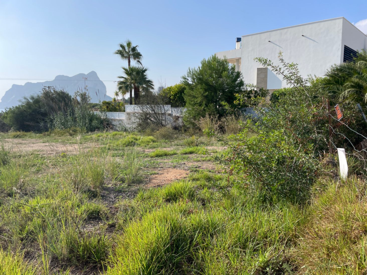 Ebenes Baugrundstück in Bassetes, Calpe, mit Blick auf den Peñón de Ifach. Das Areal ist von mediterraner Vegetation und einer modernen Villa begrenzt.