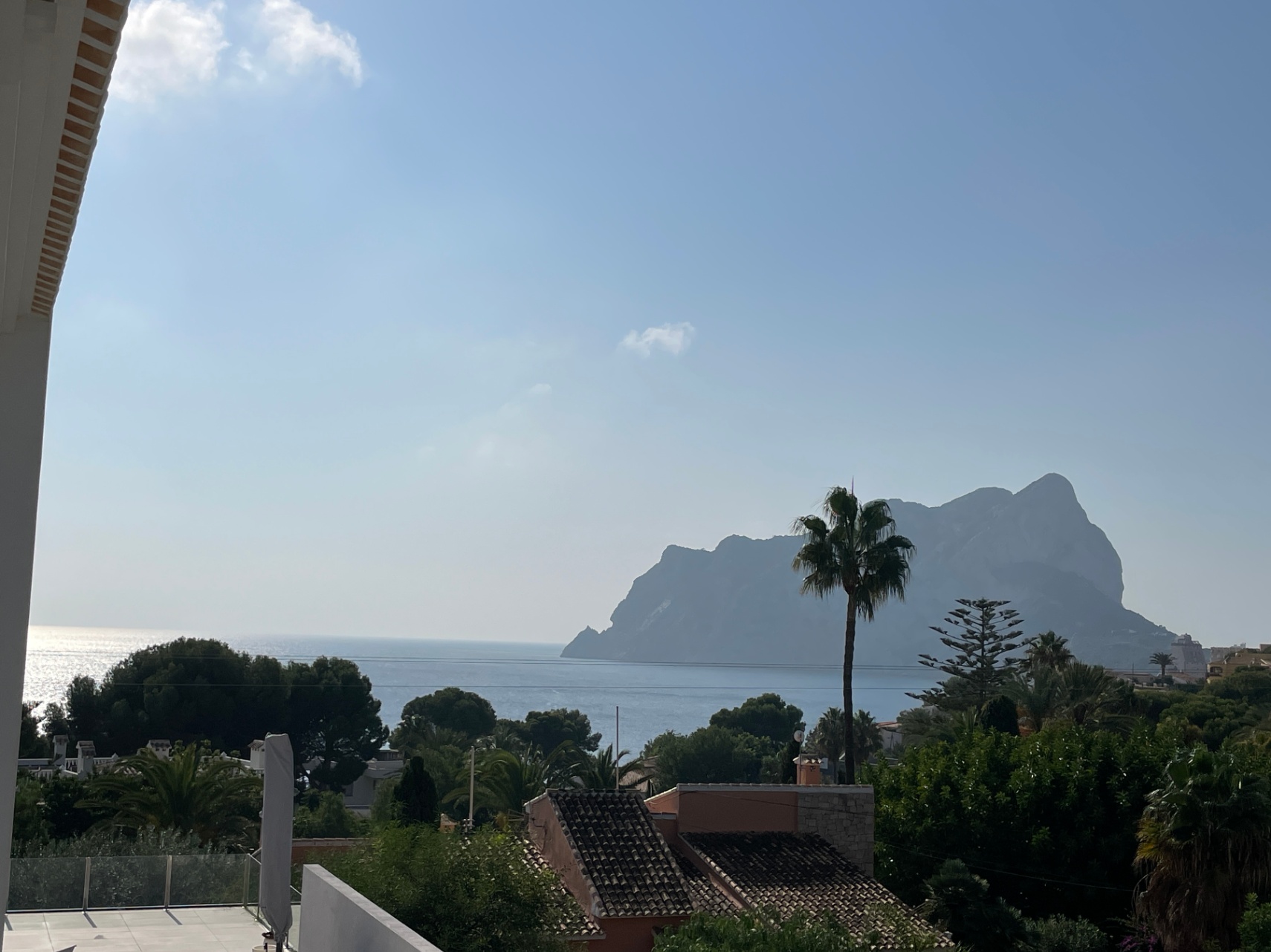 Panoramablick auf das Mittelmeer und den Peñón de Ifach von einer erhöhten Terrasse mit Glasgeländern und üppiger mediterraner Vegetation.