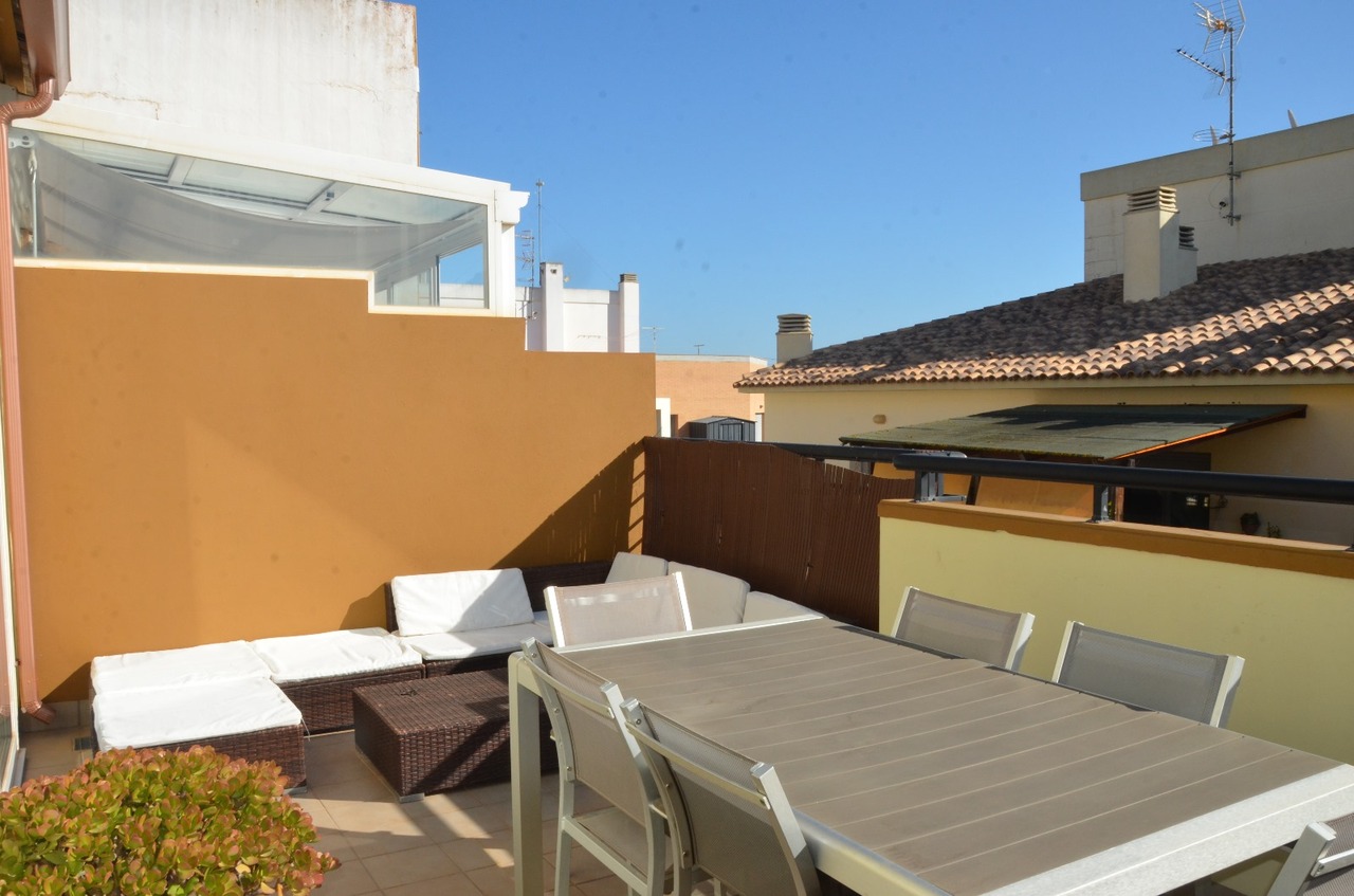 Dachterrasse mit grauem Essbereich und einer Lounge-Ecke aus Rattan mit weißen Polstern vor ockerfarbenen Wänden unter blauem Himmel.