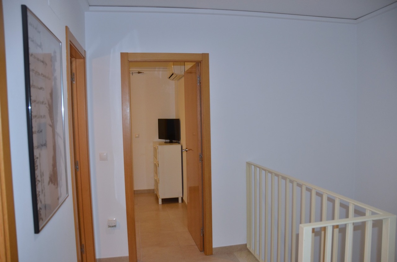Flur im Obergeschoss mit hellen Holztürrahmen, weißem Metallgeländer und Blick in ein Schlafzimmer mit weißer Kommode und Fernseher.