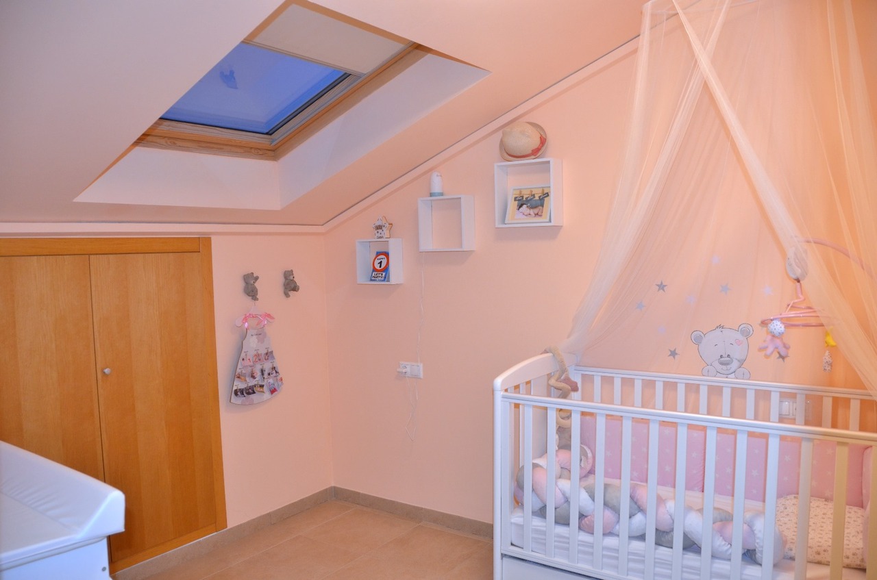 Kinderzimmer mit Dachschräge und Dachfenster, weißem Gitterbett mit Betthimmel, rosa Wänden und eingebautem Holzschrank.
