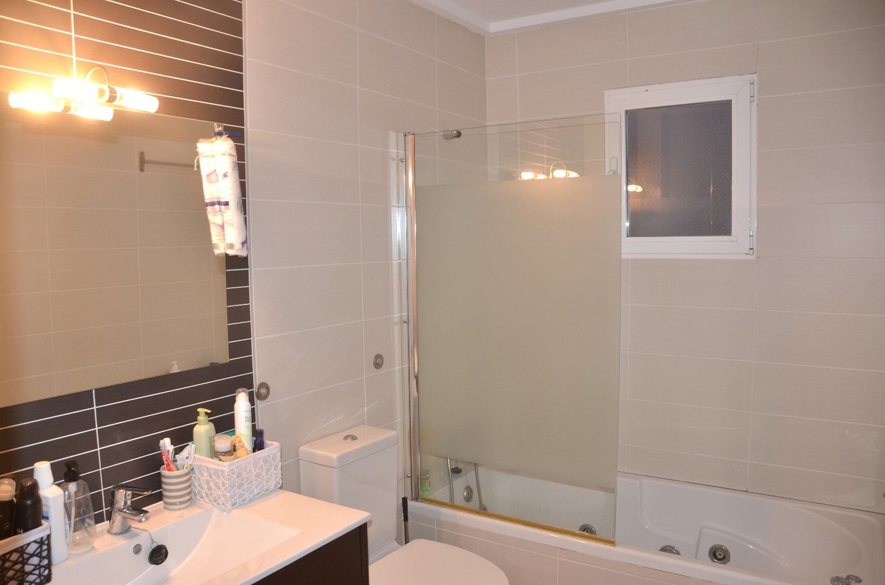Modernes Badezimmer mit weißem Waschtisch, Badewanne mit mattierter Glastrennwand und beigen Fliesen, ergänzt durch eine dunkle Akzentwand hinter dem Spiegel.