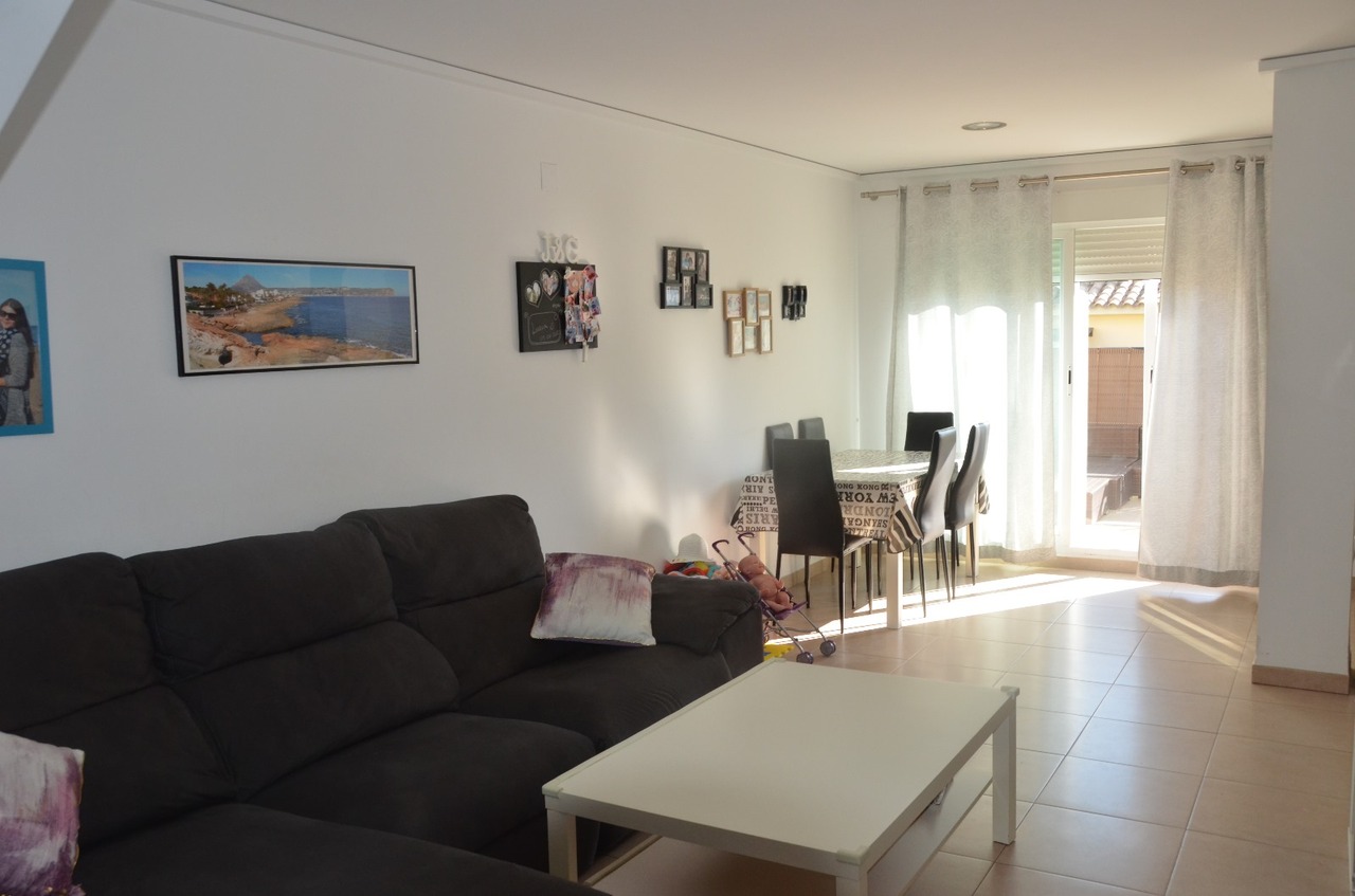 Offener Wohn- und Essbereich mit dunklem Ecksofa, weißem Couchtisch und Fliesenboden. Große Glasschiebetüren mit hellen Vorhängen führen direkt zum Außenbereich.