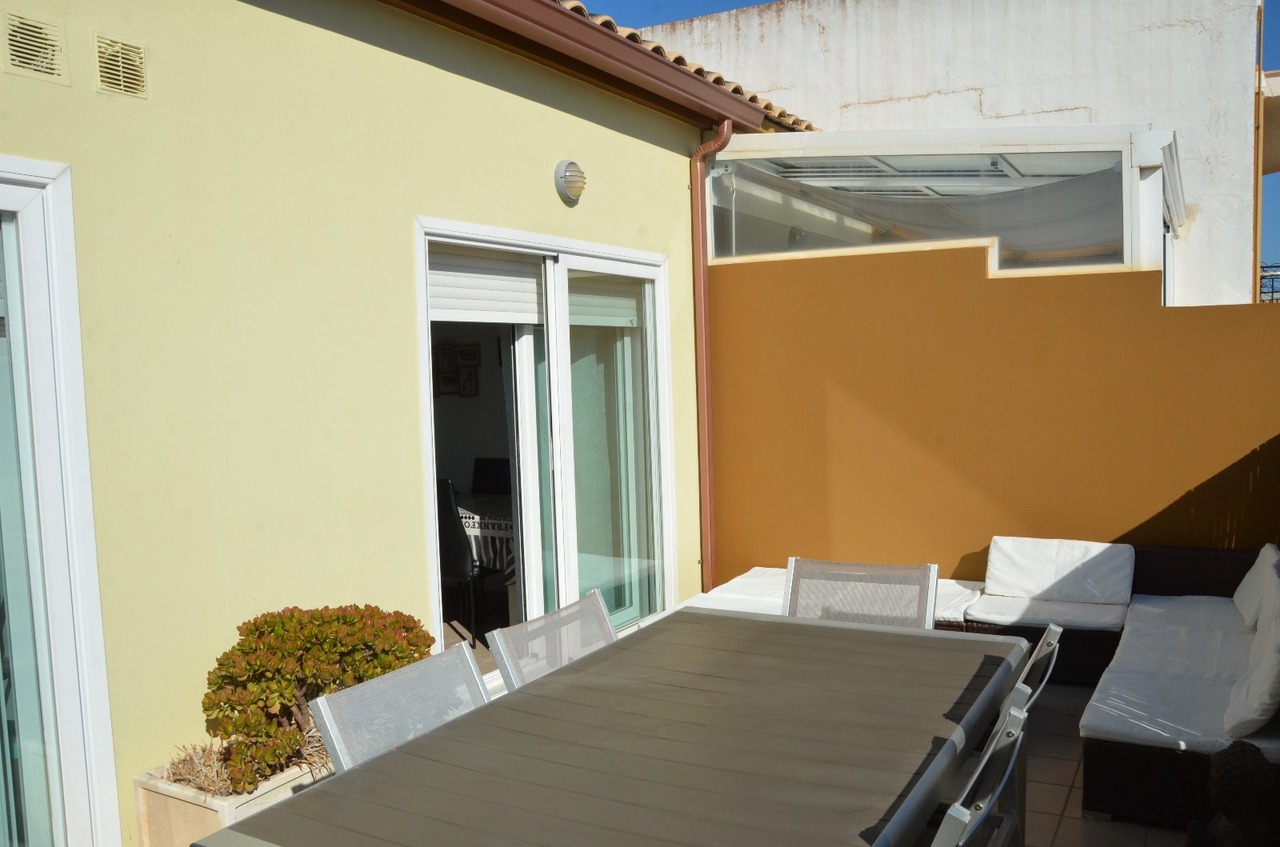 Private Terrasse mit großem Esstisch, Mesh-Stühlen und einem Loungebereich mit weißen Polstern vor einer ockerfarbenen Sichtschutzmauer.