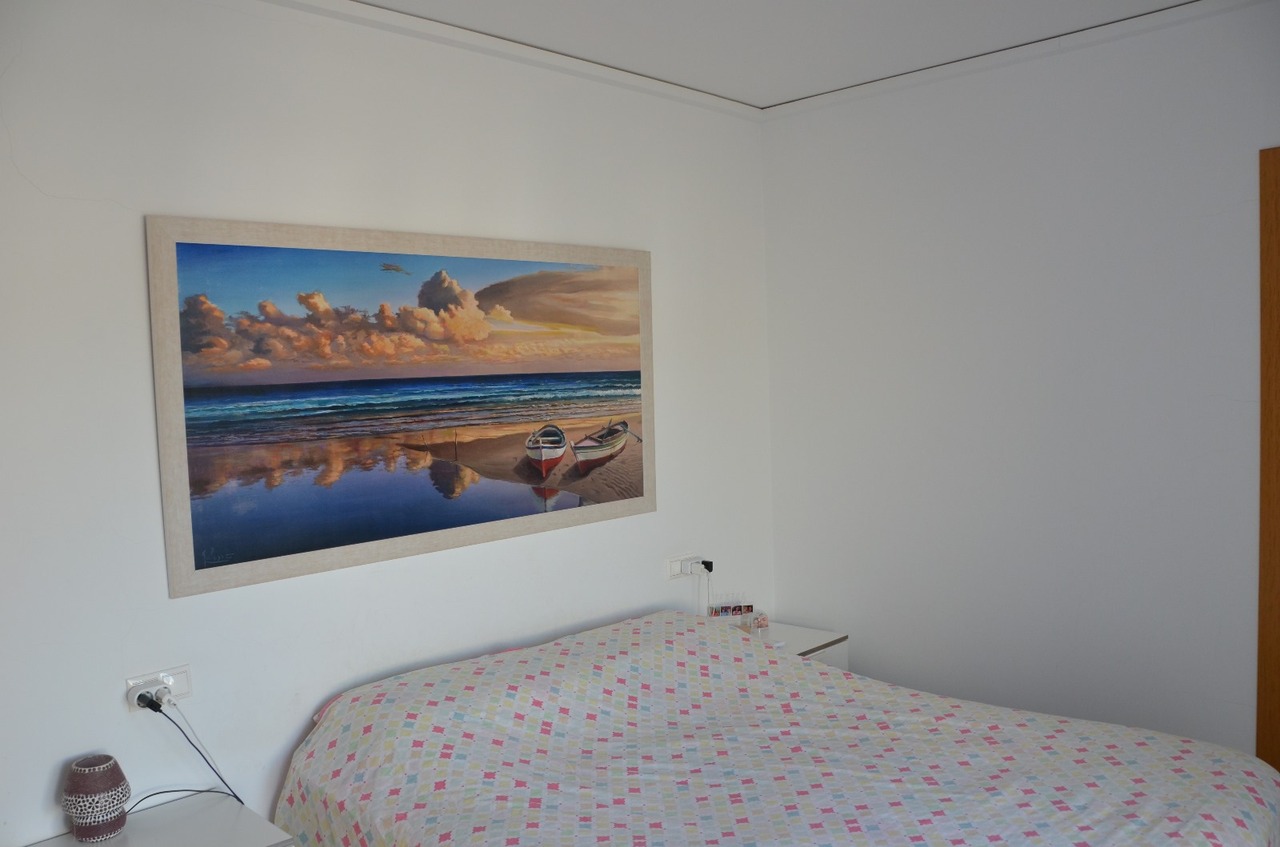 Schlafzimmer mit einem großen Küstenlandschaftsgemälde über dem Bett, weißen Wänden und einer bunt geometrisch gemusterten Tagesdecke.