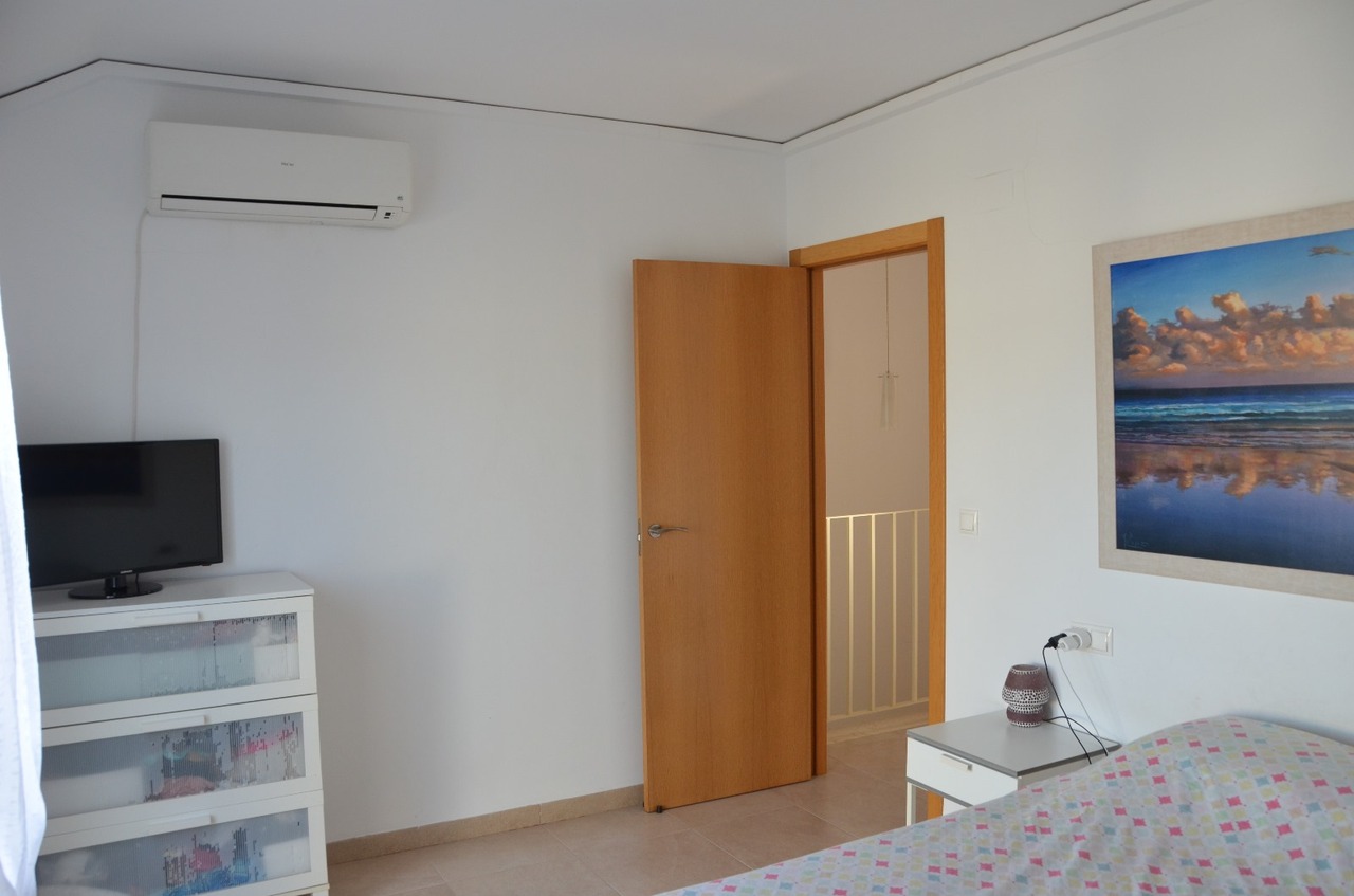 Schlafzimmer mit Split-Klimaanlage, weißer Kommode inklusive Fernseher und einer Holztür zum Flur. Ausgestattet mit Fliesenboden und dezenter Wandkunst.