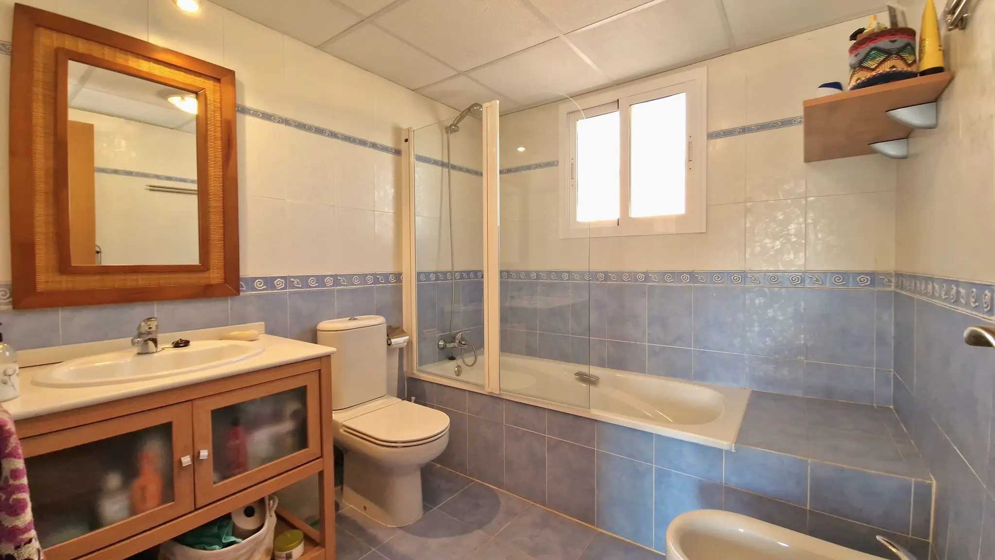 Badezimmer mit blau-weißen Fliesen, einem Holzwaschtisch und einer Badewanne mit Glasduschwand, ergänzt durch natürliches Licht durch ein Fenster.