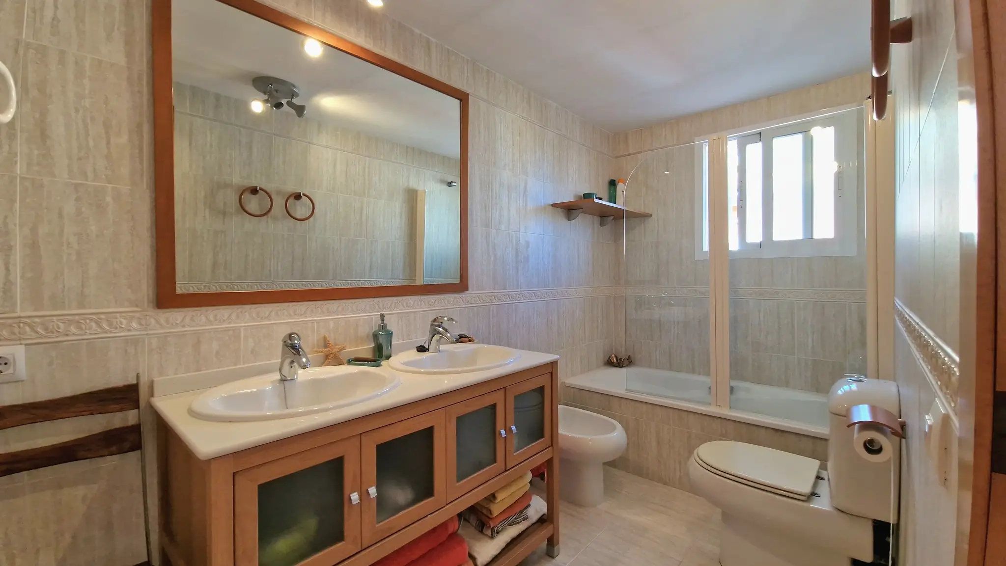 Badezimmer mit Doppelwaschbecken auf Holzunterschrank, großem Spiegel, Bidet und WC. Ausgestattet mit einer Badewanne mit Glasabtrennung und hellen Wandfliesen.