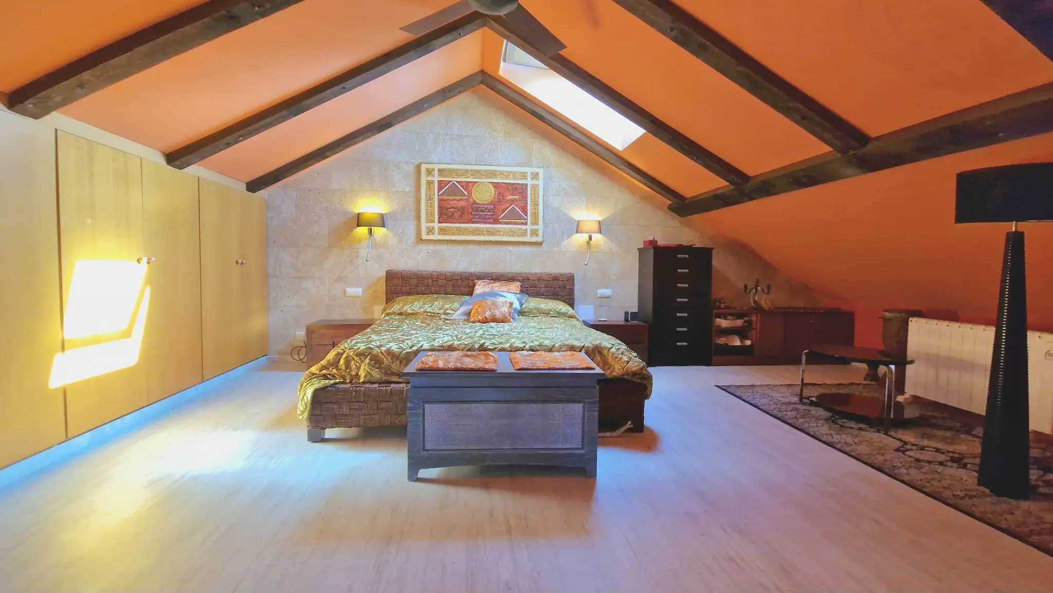 Dachgeschoss-Schlafzimmer mit dunklen Holzbalken, orangefarbenen Deckenschrägen, einer Steinakzentwand und hellen Einbauschränken aus Holz.