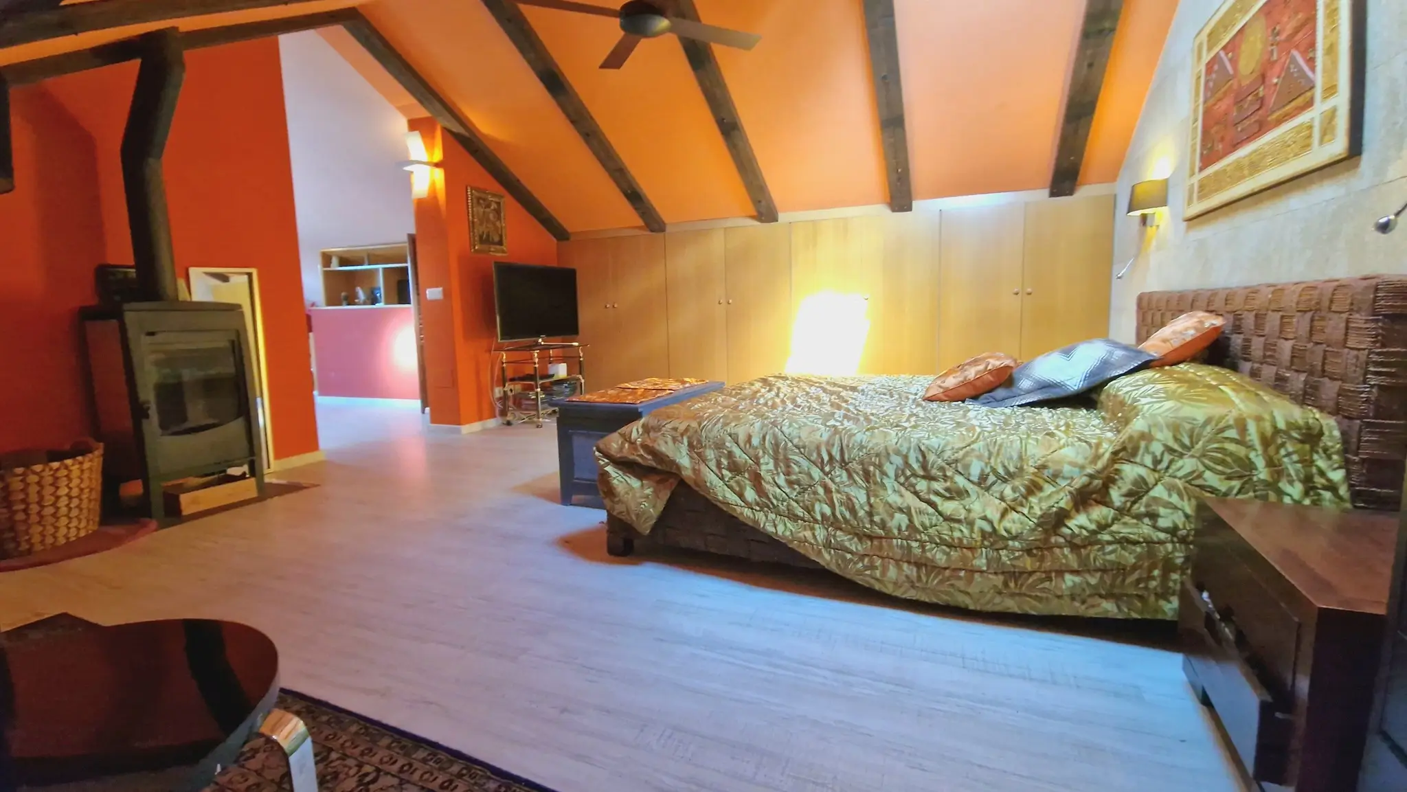 Dachgeschoss-Schlafzimmer mit sichtbaren Holzbalken, Kaminofen und terracottafarbenen Wänden. Mit Einbauschränken, Deckenventilator und geflochtenem Kopfteil.