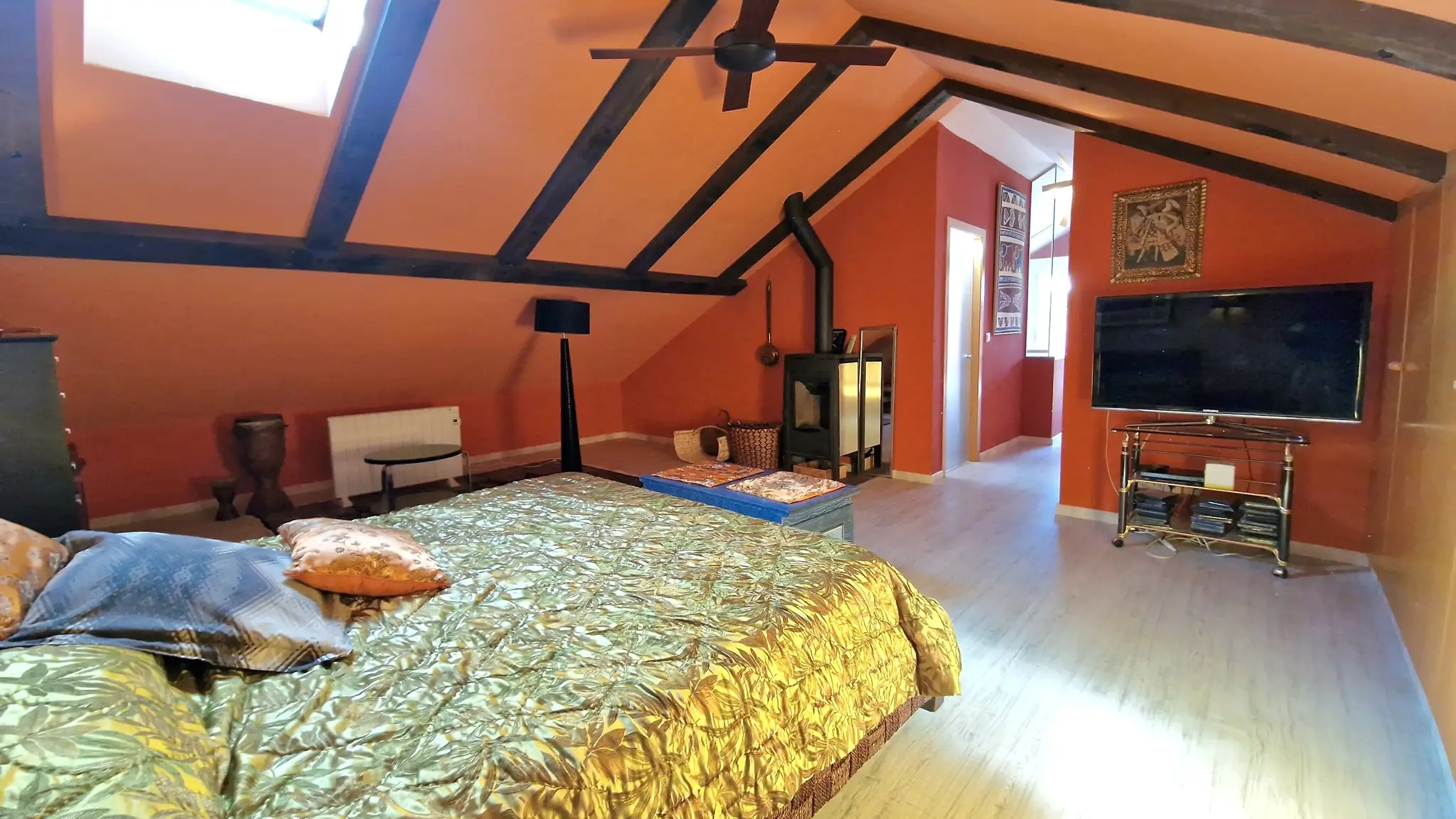 Dachgeschoss-Schlafzimmer mit sichtbaren Holzbalken, terracottafarbenen Wänden, einem Kaminofen und einem Dachfenster für natürliches Licht.