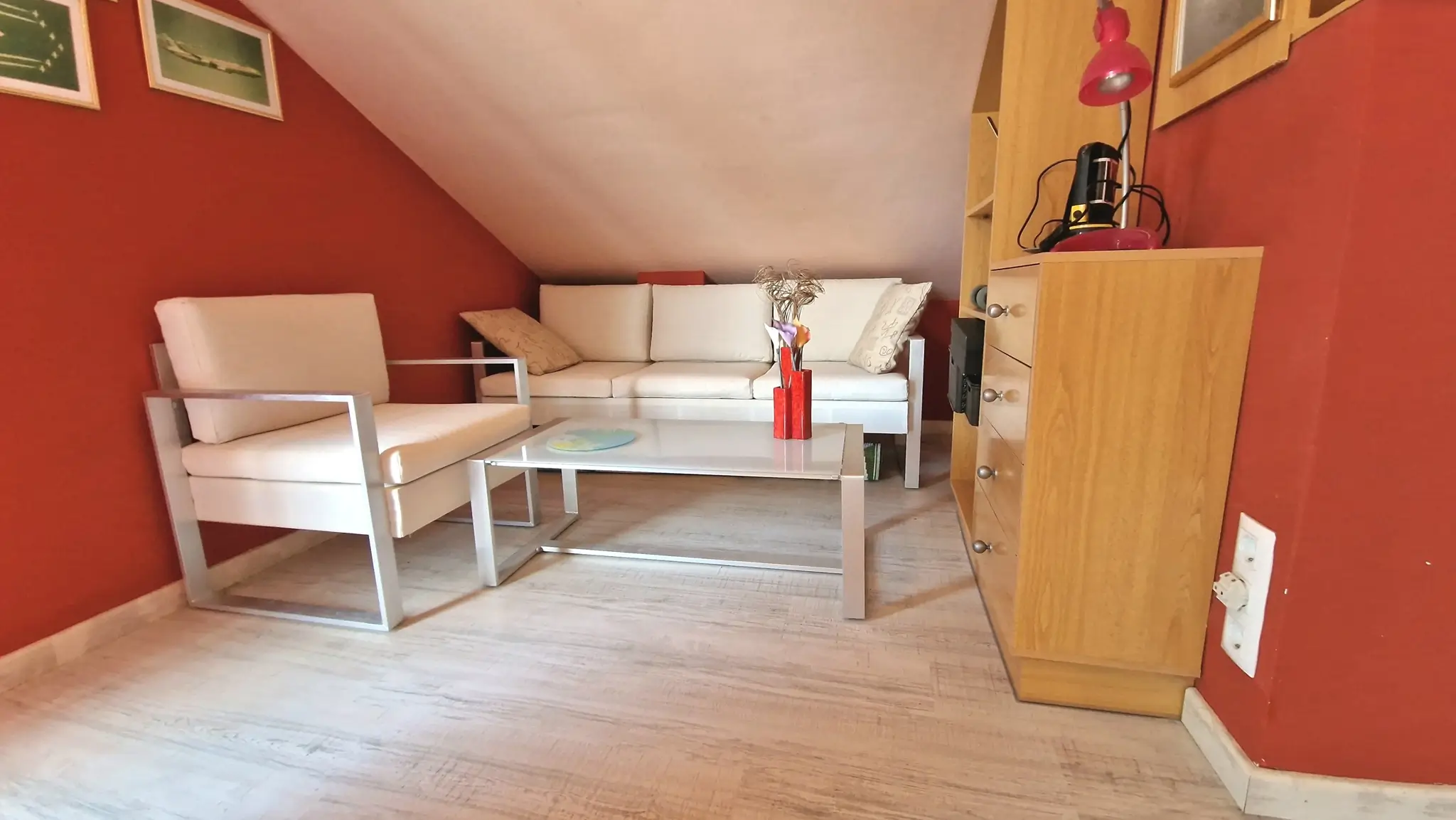 Dachwohnzimmer mit roten Wänden, weißen Metallrahmen-Sitzmöbeln, Glastisch und hellem Bodenbelag in Holzoptik.