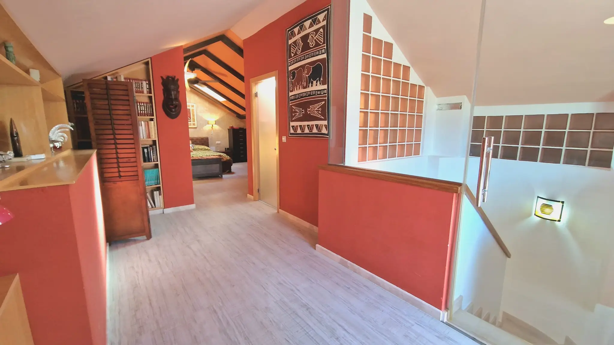 Flur im Obergeschoss mit terracottafarbenen Wänden und hellem Holzboden. Der Bereich bietet Zugang zu einem Schlafzimmer mit sichtbaren Dachbalken und integrierten Holzregalen.