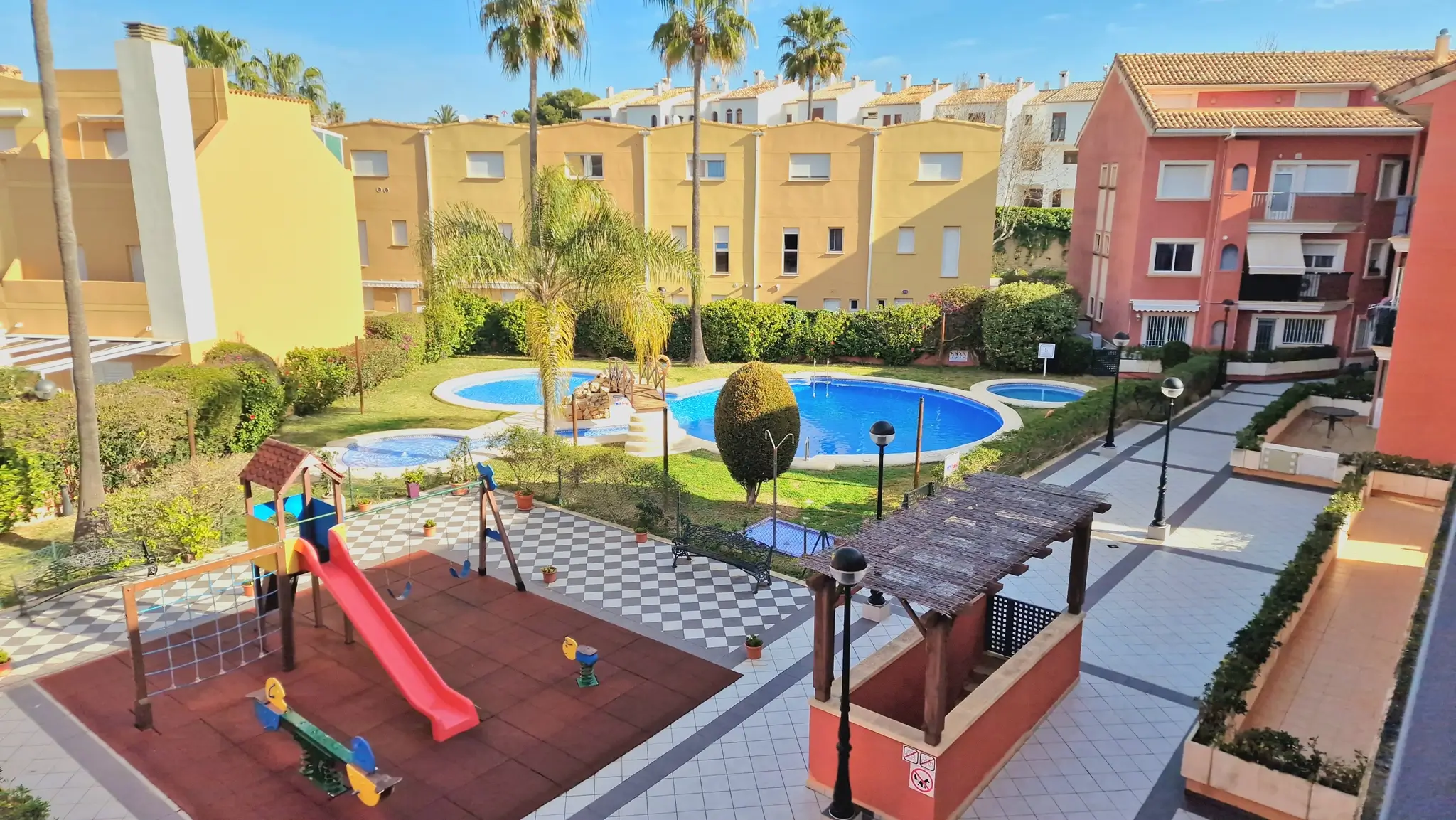 Gemeinschaftsanlage mit Kinderspielplatz, großem Swimmingpool mit Brücke und gepflegten Gärten, eingebettet in eine mediterrane Wohnanlage mit Palmen.