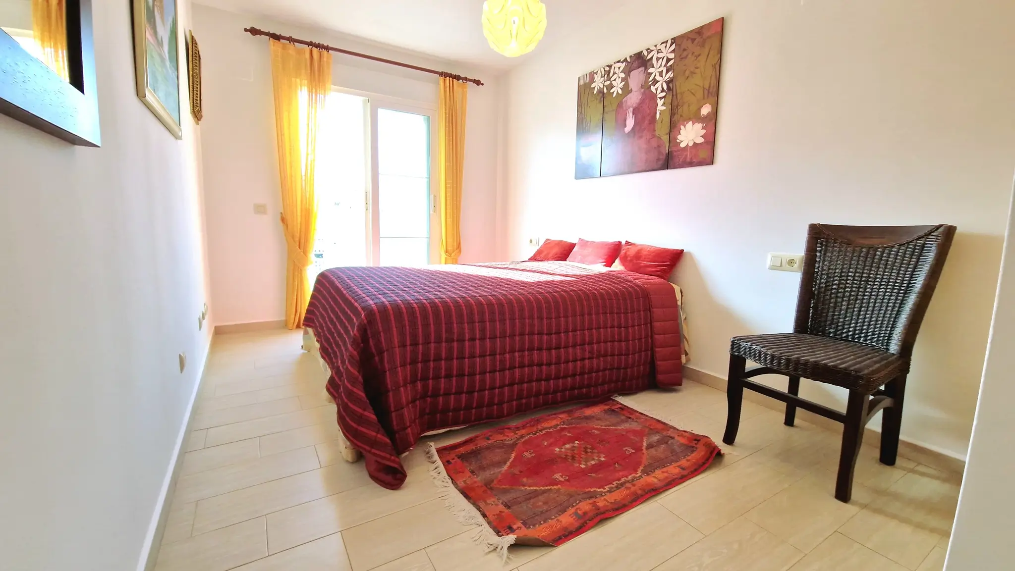 Helles Schlafzimmer mit Doppelbett, roter Steppdecke, gelben Vorhängen, einem dunklen Korbstuhl und einem dekorativen Buddha-Triptychon an der Wand.