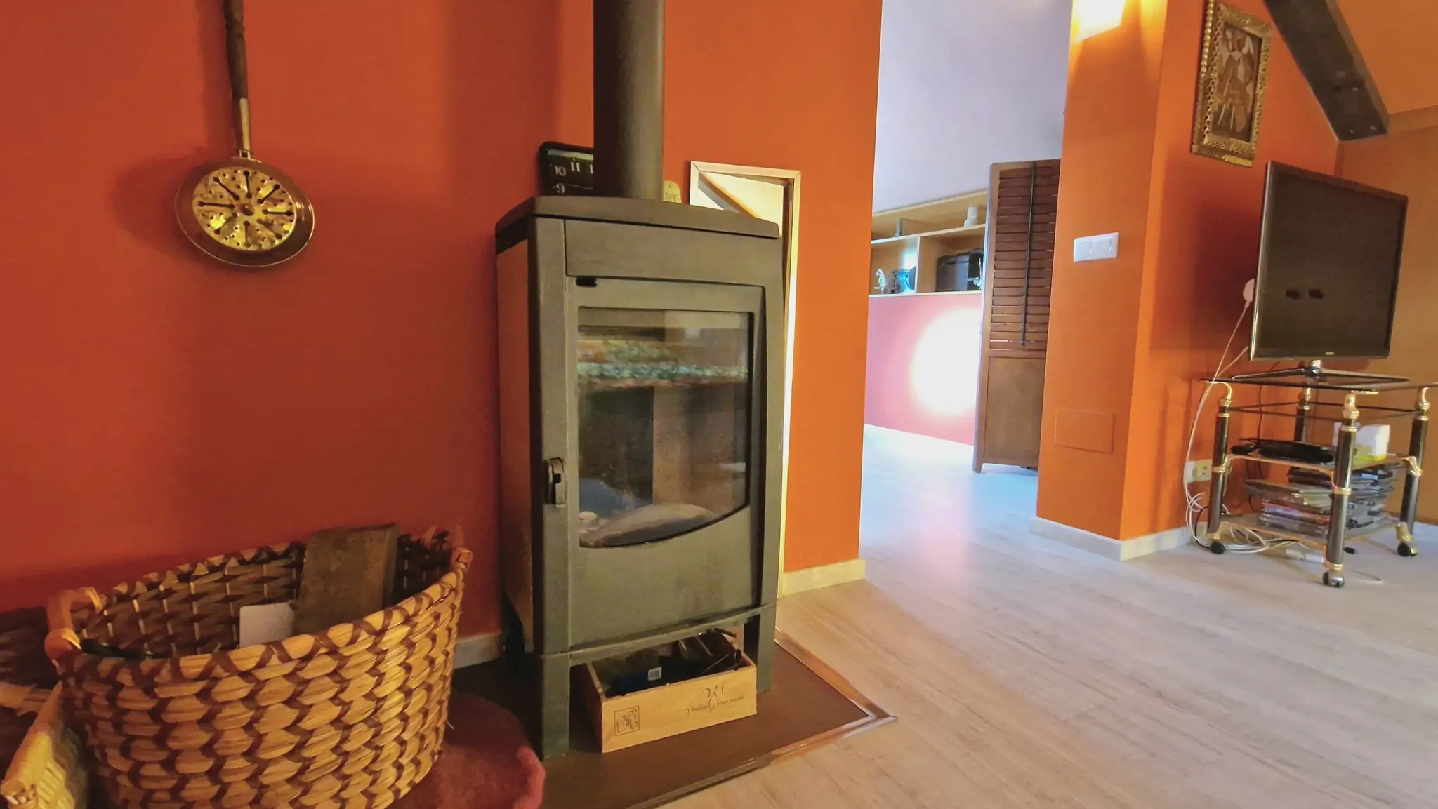 Moderner schwarzer Kaminofen vor terrakottafarbenen Wänden, kombiniert mit hellem Holzboden und einem traditionellen Weidenkorb für Kaminholz.