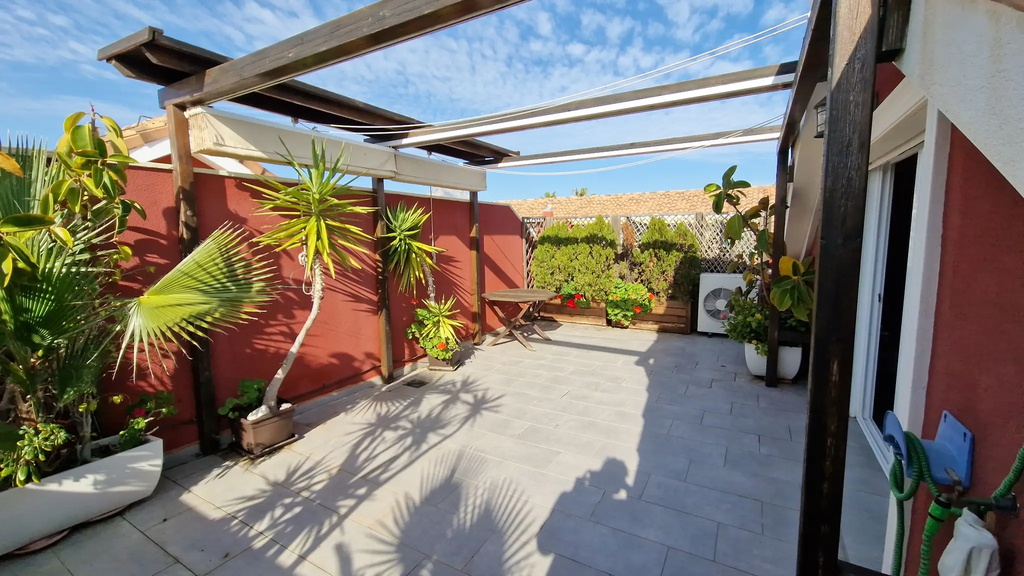 Private Terrasse mit grauen Fliesen, dunkler Holzpergola und ausziehbaren Markisen. Terrakotta-Wände, Palmen in Kübeln und ein runder Holztisch unter freiem Himmel.