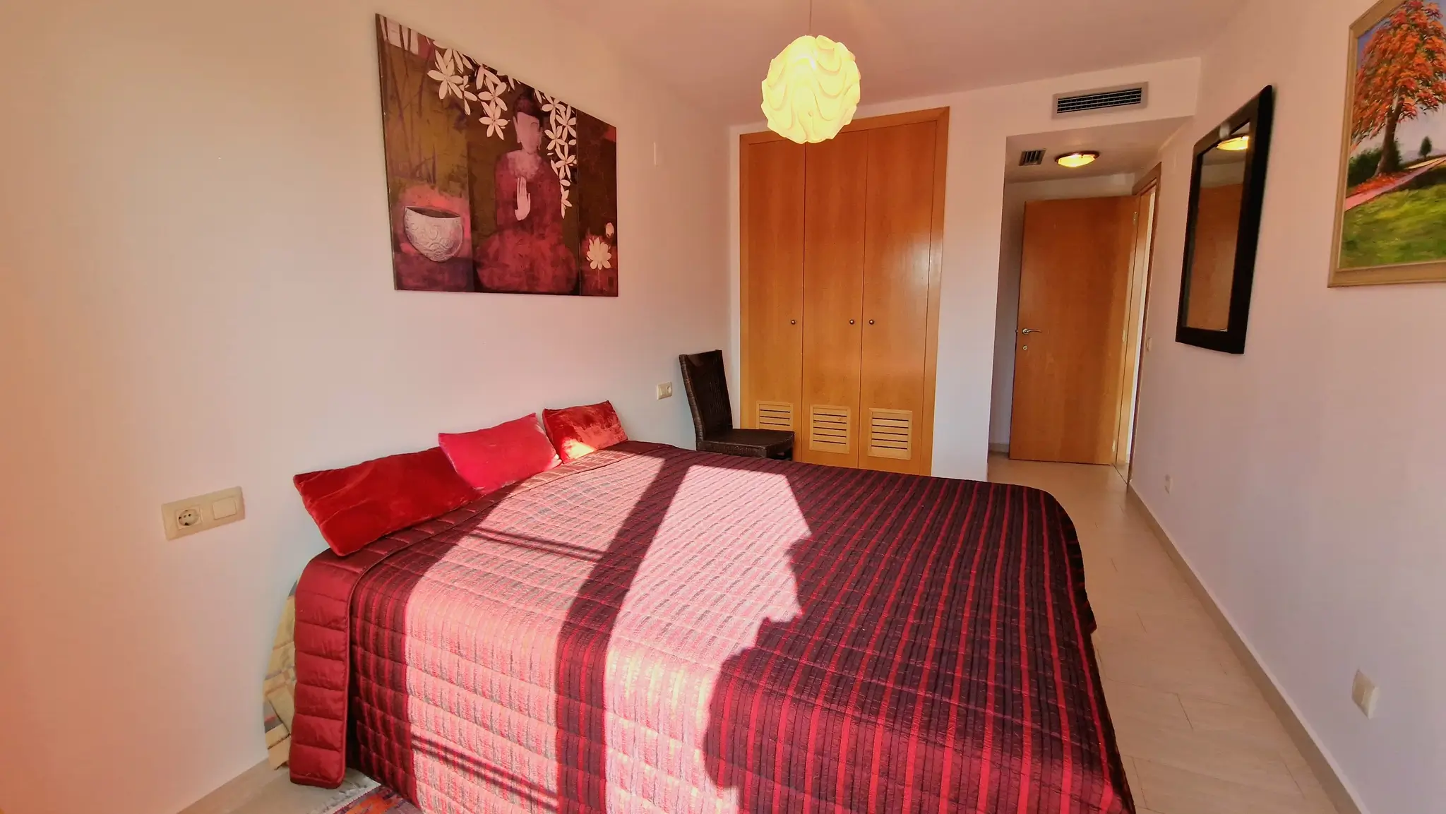 Schlafzimmer mit Doppelbett, weinroter gestreifter Tagesdecke, Einbauschränken aus Holz und Wandkunst mit Buddha-Motiv.