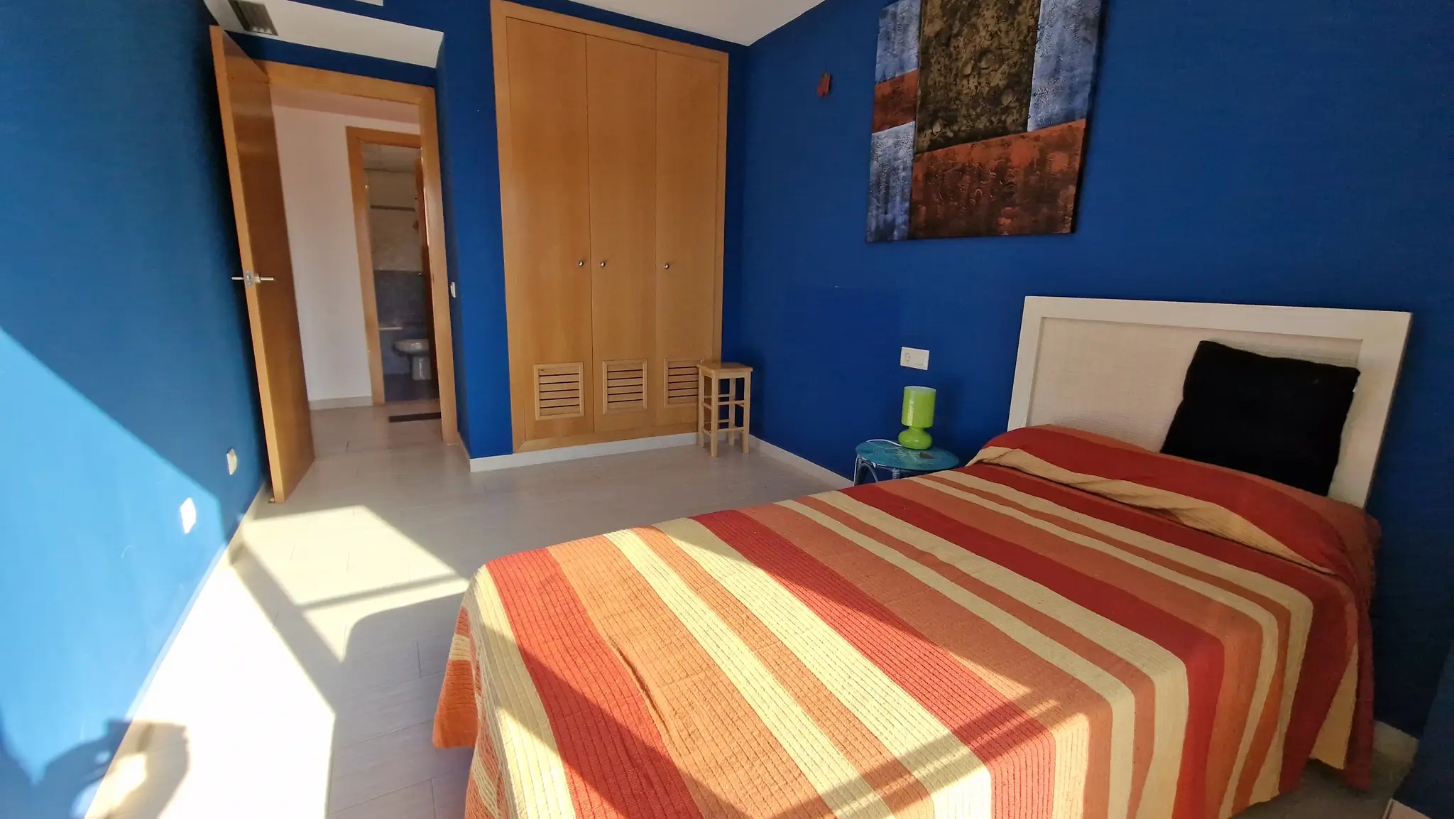 Schlafzimmer mit tiefblauen Wänden, Einbauschrank aus Holz und Fliesenboden. Der Raum verfügt über ein Einzelbett und direkten Zugang zu einem angrenzenden Badezimmer.