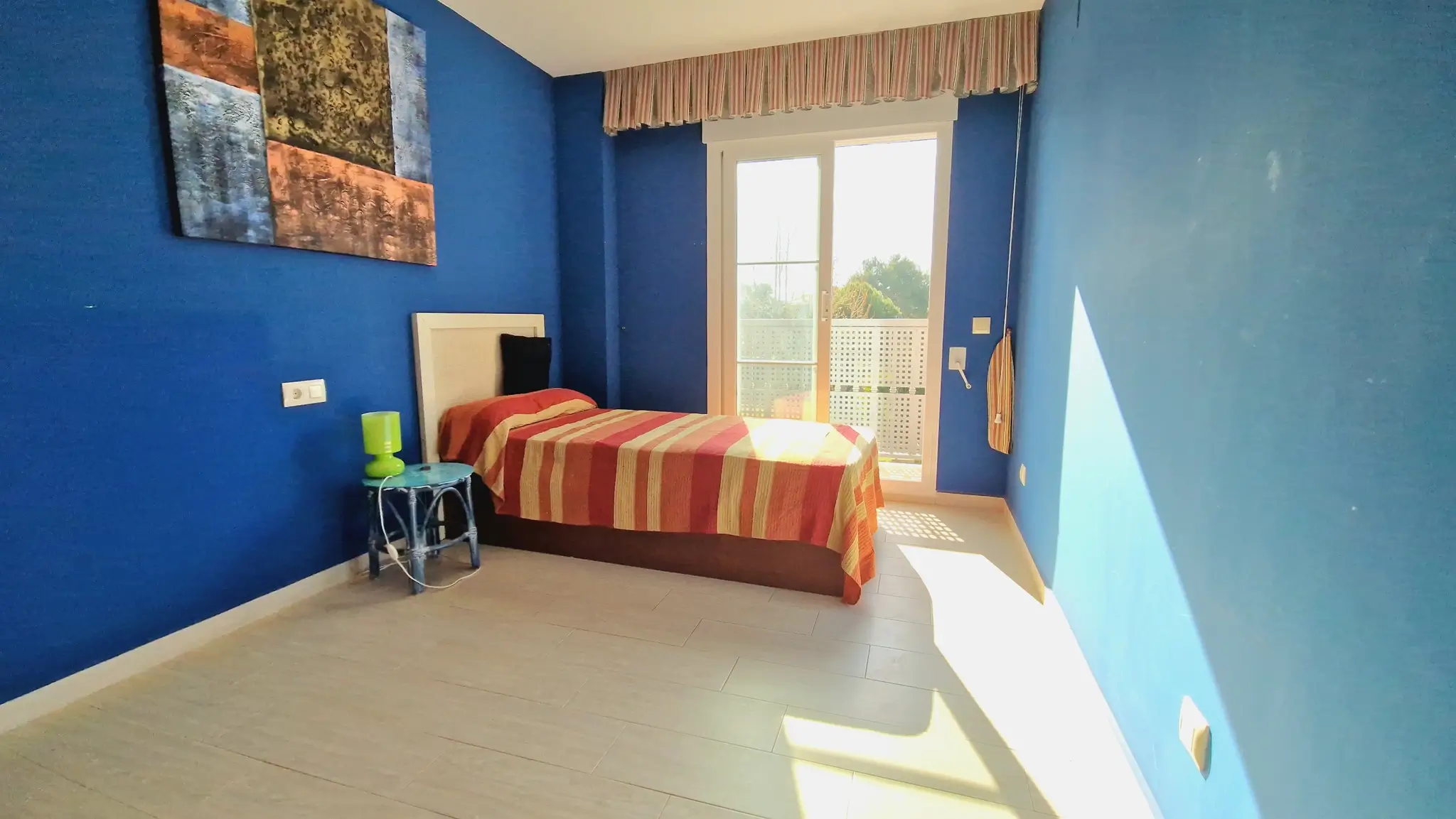 Schlafzimmer mit tiefblauen Wänden, Einzelbett mit gestreifter Bettwäsche und direktem Zugang zum Balkon durch Glasschiebetüren.