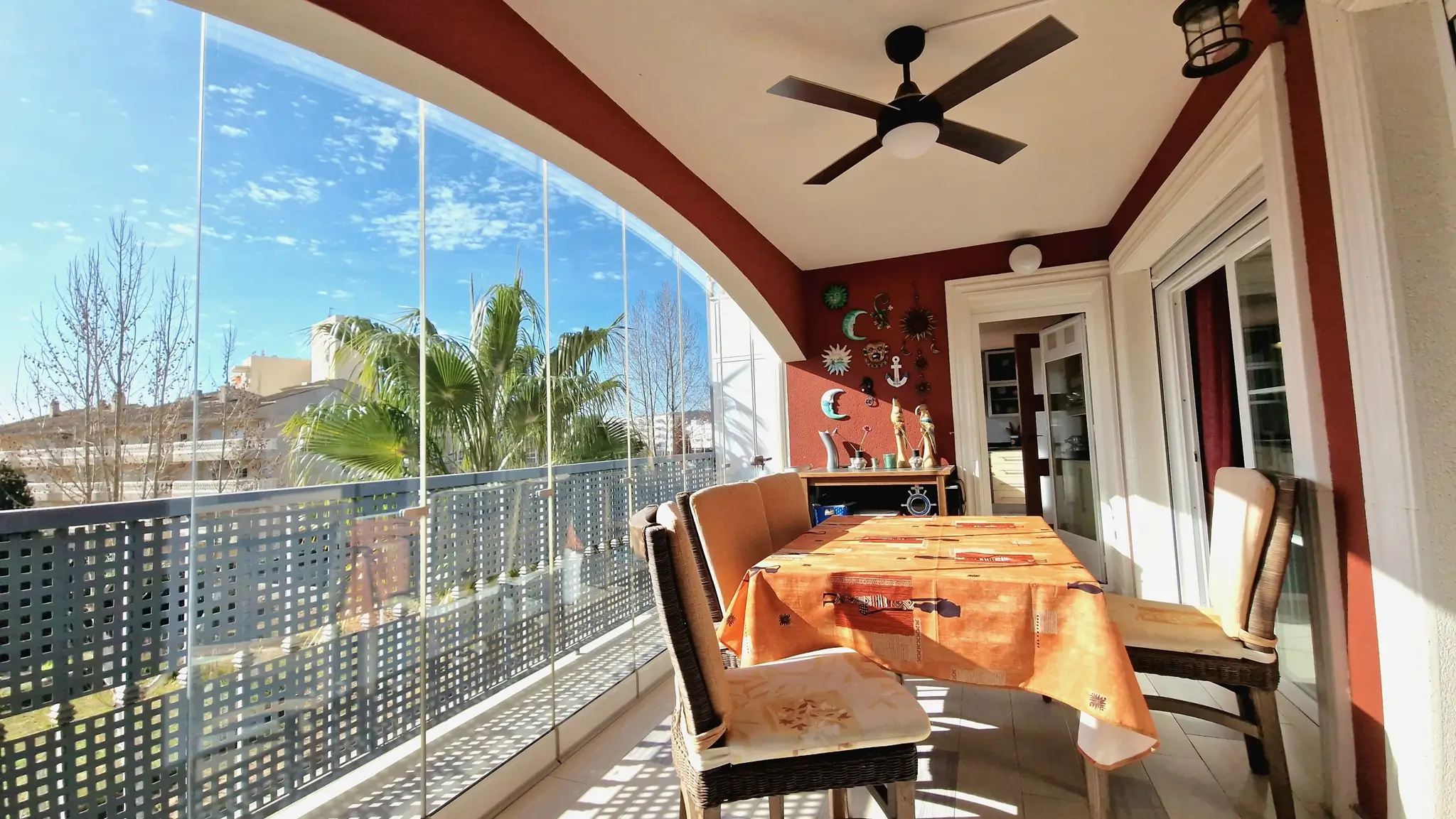 Überdachte Terrasse mit rahmenlosen Glasvorhängen, Essbereich im Freien mit Rattanstühlen, Deckenventilator und Blick auf Palmen und blauen Himmel.