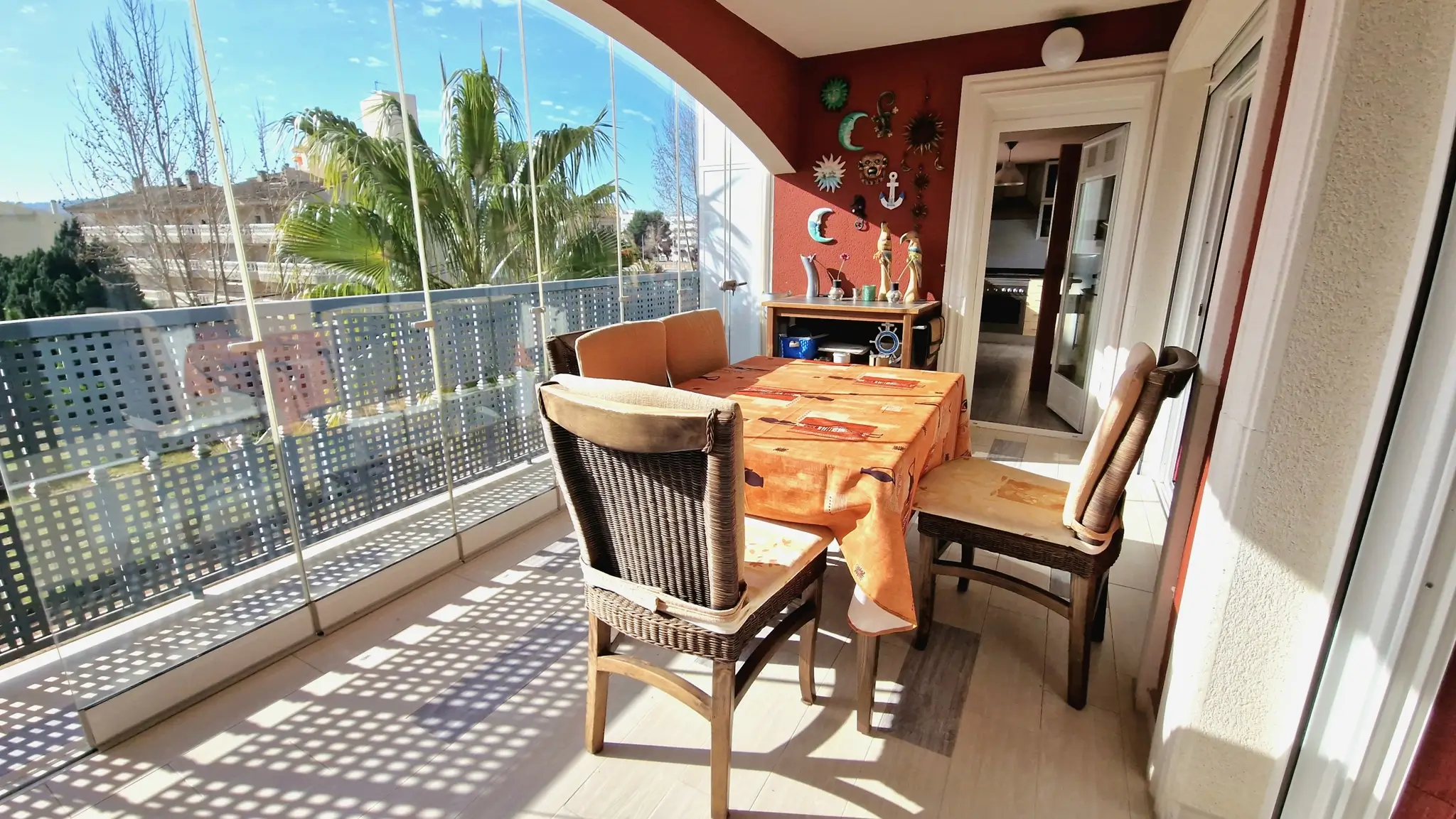 Verglaste Terrasse mit Glasschiebewänden, Essbereich mit Korbstühlen und einer roten Akzentwand mit Dekoration und Blick auf Palmen.