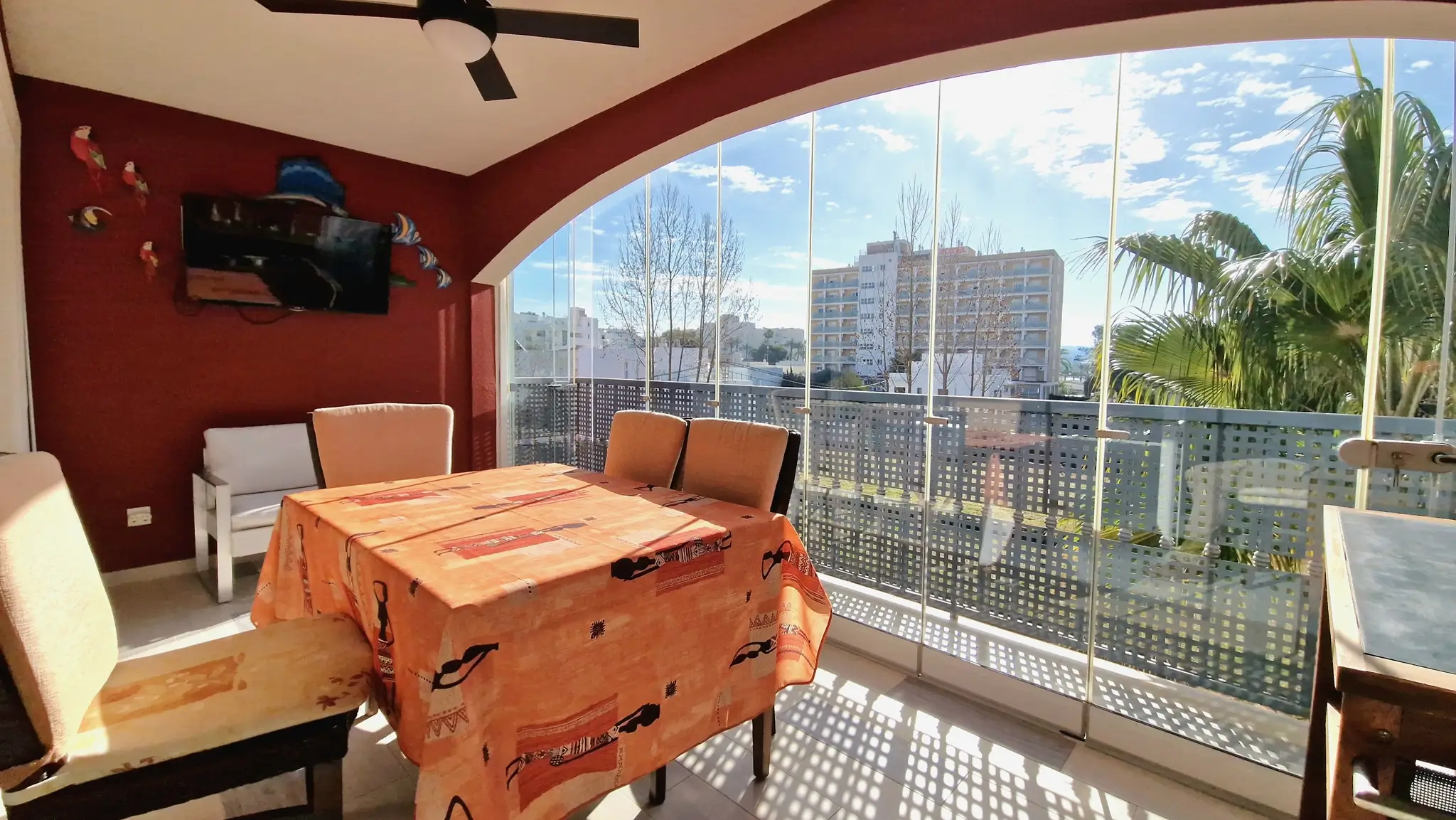 Verglaste Terrasse mit rahmenlosen Glasschiebewänden, Essbereich, roter Akzentwand mit Fernseher und Blick auf die städtische Umgebung.