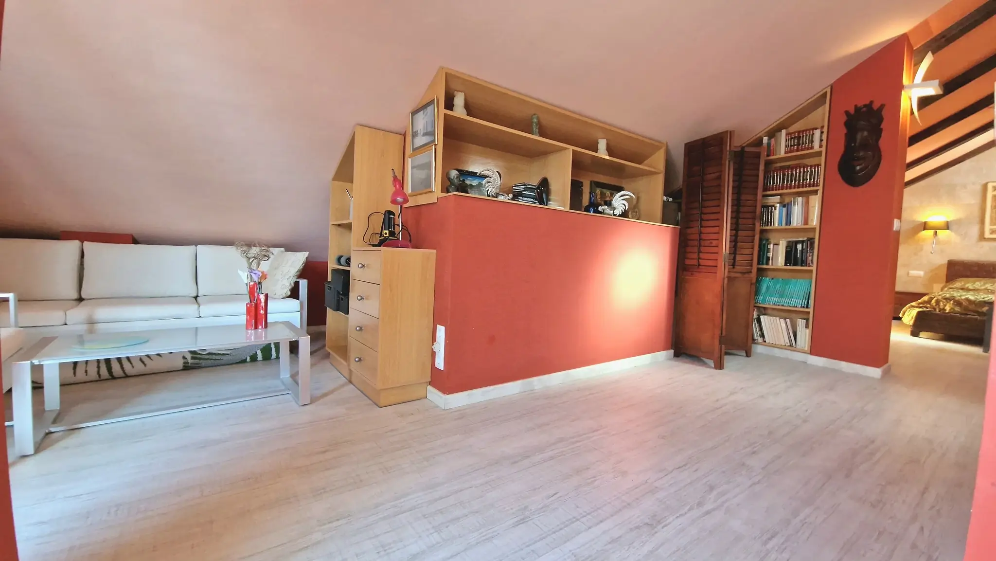 Wohnbereich im Dachgeschoss mit terrakottafarbenen Akzentwänden, integrierten Holzregalen und hellem Laminatboden unter einer Dachschräge.
