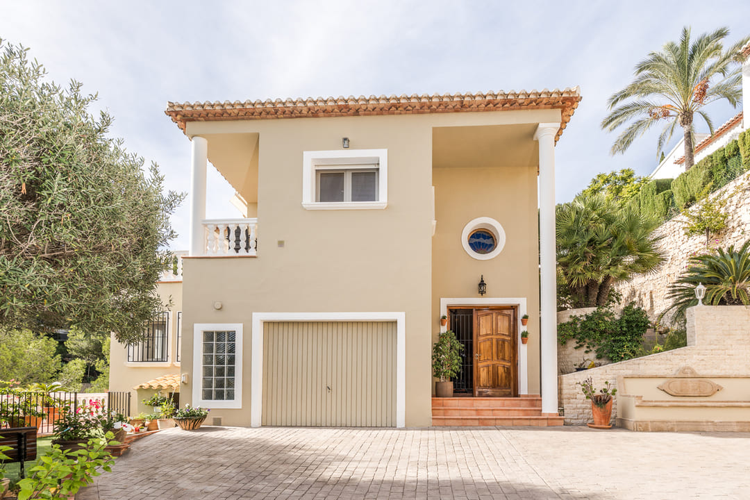 Außenfassade einer zweistöckigen Villa mit beigem Putz, integrierter Garage, Holzeingangstür und gepflasterter Einfahrt inmitten mediterraner Bepflanzung.