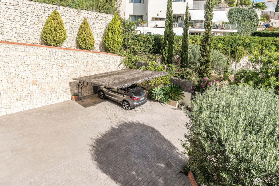 Außenparkplatz mit gepflasterter Einfahrt und Heidekraut-Carport, begrenzt durch eine Natursteinmauer und gepflegte mediterrane Bepflanzung.
