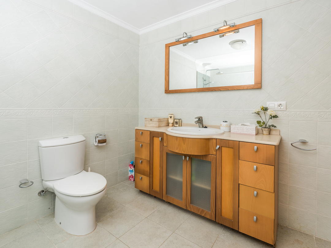 Badezimmer mit einem Waschtisch aus Holz, integriertem Waschbecken, großem Spiegel mit Holzrahmen und Beleuchtung sowie hellen, gemusterten Wandfliesen.