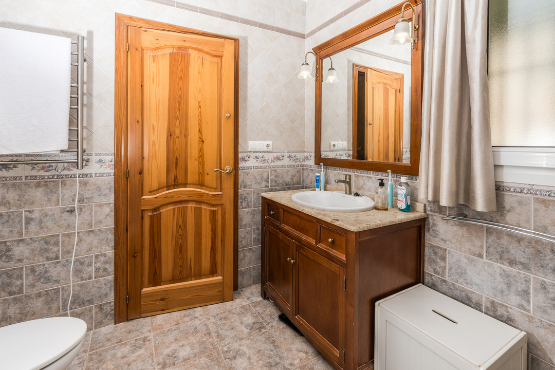 Badezimmer mit Holzwaschtisch, Steinplatte und passendem Spiegel. Wände mit dekorativen Fliesen und Blumenbordüre, Naturholztür und Handtuchheizkörper.