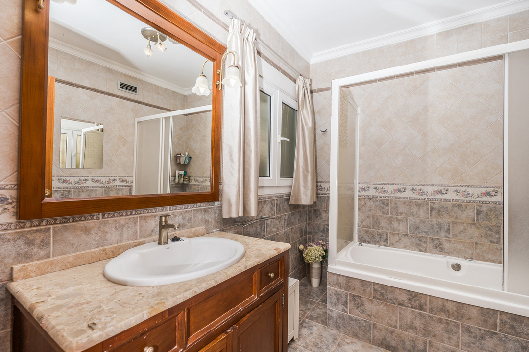 Badezimmer mit Holzwaschtisch und Marmorplatte, großem Wandspiegel mit Holzrahmen sowie einer Badewanne mit Schiebetür und dekorativen Fliesendetails.