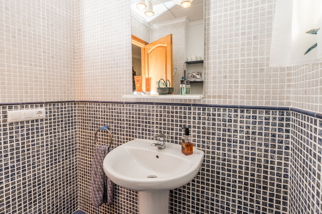 Badezimmer mit weißem Standwaschbecken, Wänden aus weiß-blauen Mosaikfliesen und einem Spiegel mit integrierter Beleuchtung und Ablage.