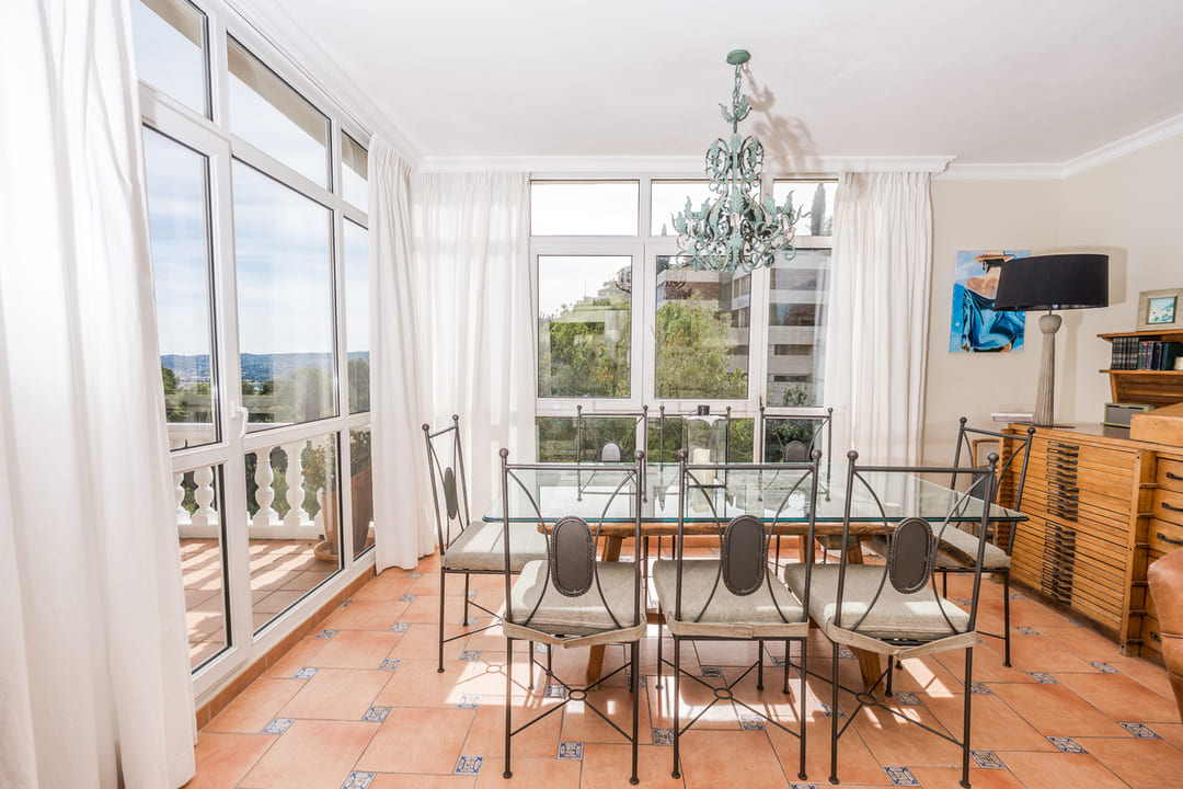 Esszimmer mit Glastisch und schmiedeeisernen Stühlen auf Terrakottafliesen. Große Fensterfronten sorgen für viel Licht und bieten einen weiten Blick ins Grüne.
