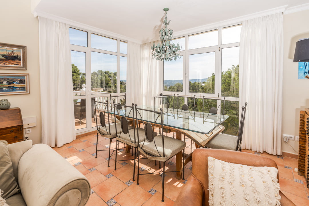 Heller Essbereich mit Glastisch, schmiedeeisernen Stühlen und Terrakotta-Fliesen. Große Fensterfronten bieten einen weiten Blick auf die grüne Landschaft.