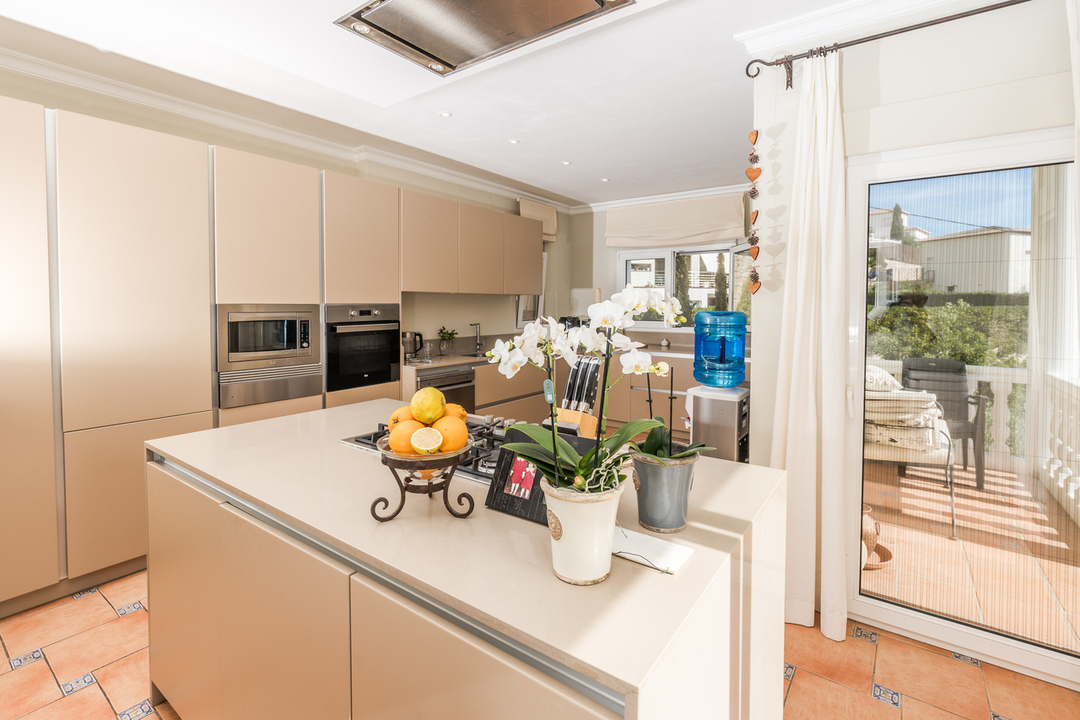 Moderne Küche mit beiger Kochinsel, Gaskochfeld, integrierten Geräten und Terrakottaboden. Eine große Glastür führt direkt auf die angrenzende Terrasse.