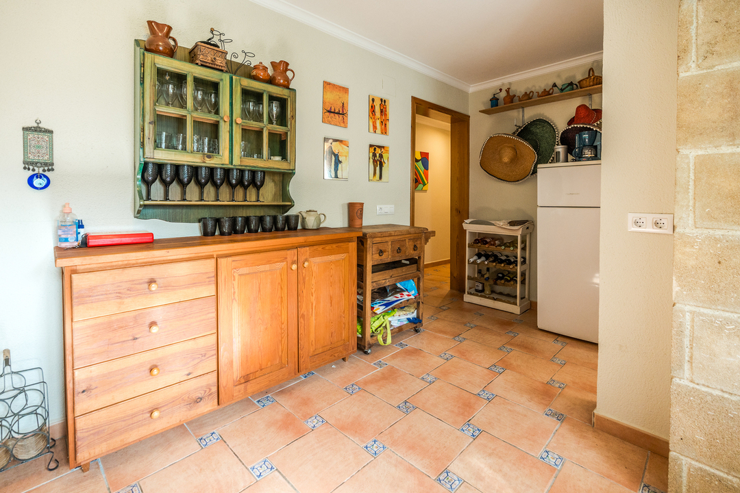 Rustikale Holzkommode und grüner Hängeschrank für Gläser über Terrakottafliesen mit dekorativen Einlagen. Ein weißer Kühlschrank und Weinregal ergänzen den funktionalen Raum.