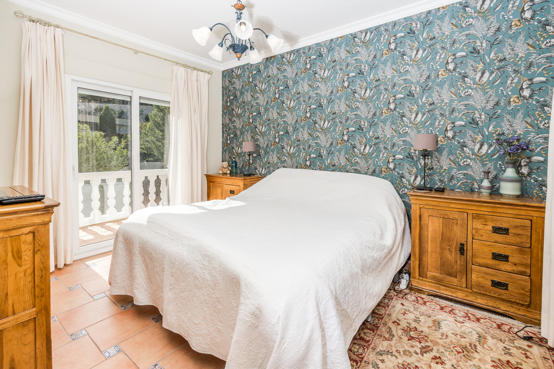 Schlafzimmer mit botanischer Mustertapete, Massivholzmöbeln und Terrakottaboden. Eine Glasschiebetür führt auf den Balkon mit weißer Balustrade.