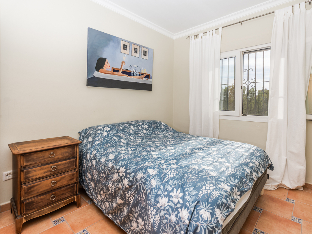 Schlafzimmer mit Doppelbett, blau-floraler Bettwäsche, klassischem Holznachtschrank und Terrakotta-Fliesenboden mit dekorativen Einlegern.
