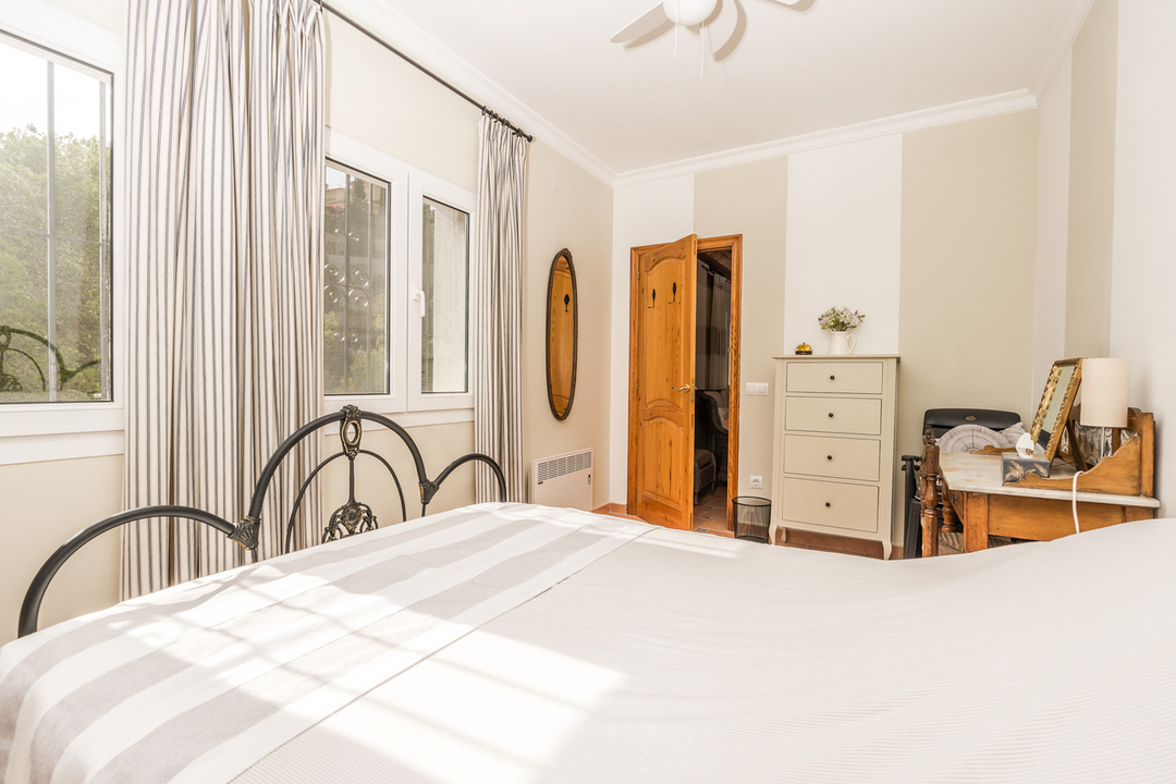 Schlafzimmer mit schmiedeeisernem Kopfteil, klassischem Holzschreibtisch und Fenstern mit gestreiften Vorhängen, die viel Tageslicht hereinlassen.