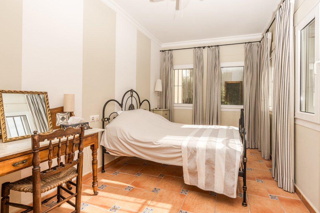 Schlafzimmer mit schwarzem Schmiedeeisenbett, gestreifter Tapete und Terrakottaboden mit dekorativen Fliesen. Ein klassischer Holzschreibtisch mit Spiegel steht daneben.