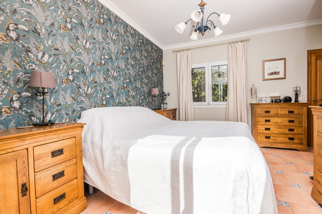 Schlafzimmer mit tropisch gemusterter Akzentwand, Massivholzkommoden und Terrakotta-Fliesenboden mit dekorativen Einlegern. Natürliches Licht flutet durch das Fenster.