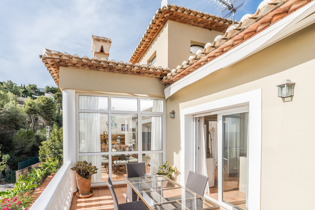 Terrasse mit Terrakottaboden, Glastisch und Korbstühlen vor weißer Balustrade. Blick auf die mediterrane Fassade, einen Wintergarten und die bewaldeten Hügel der Umgebung.