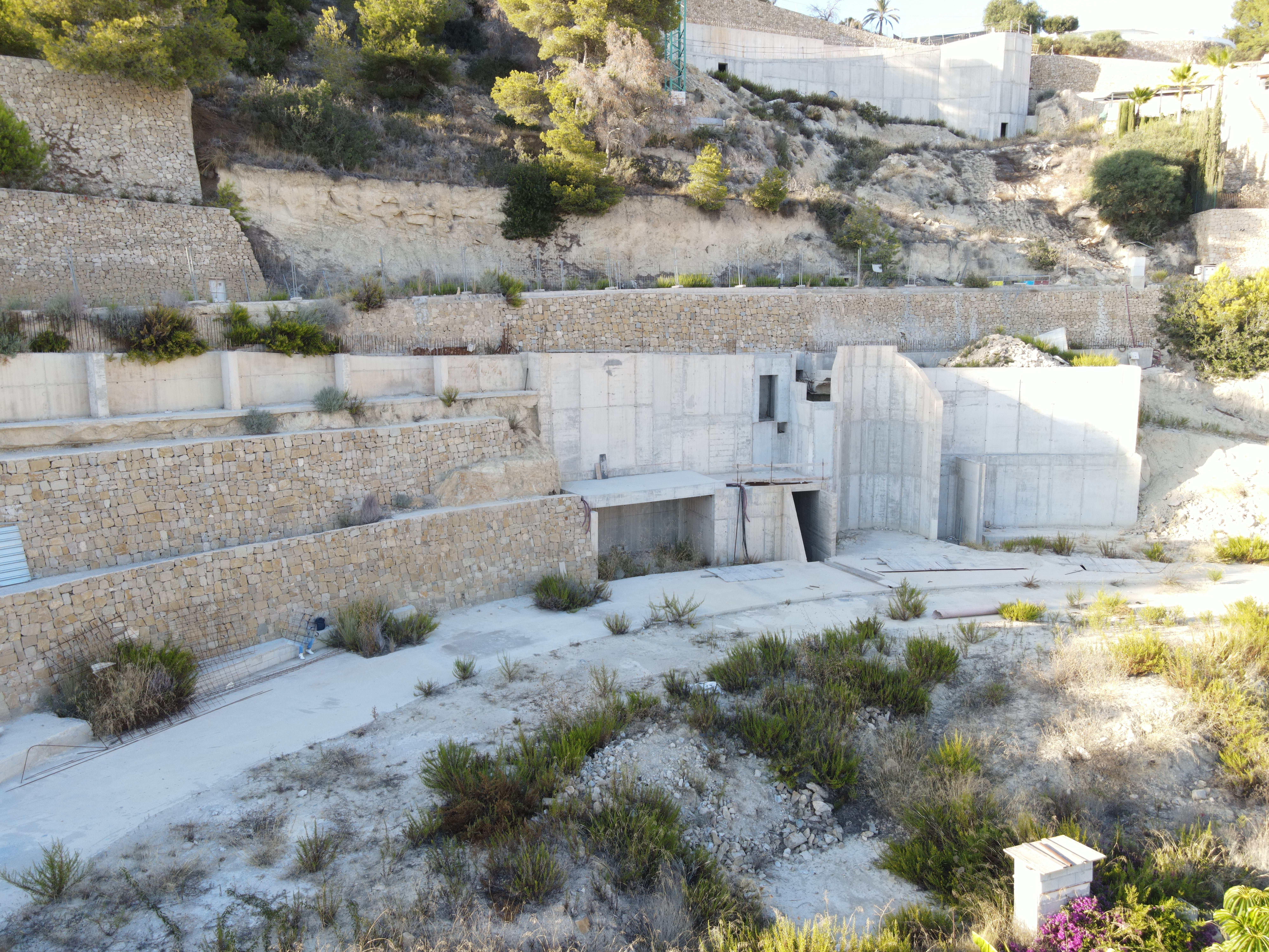 Baustellenansicht mit gestuften Trockensteinmauern und in den Hang integrierten Stahlbetonkonstruktionen sowie mediterraner Vegetation.
