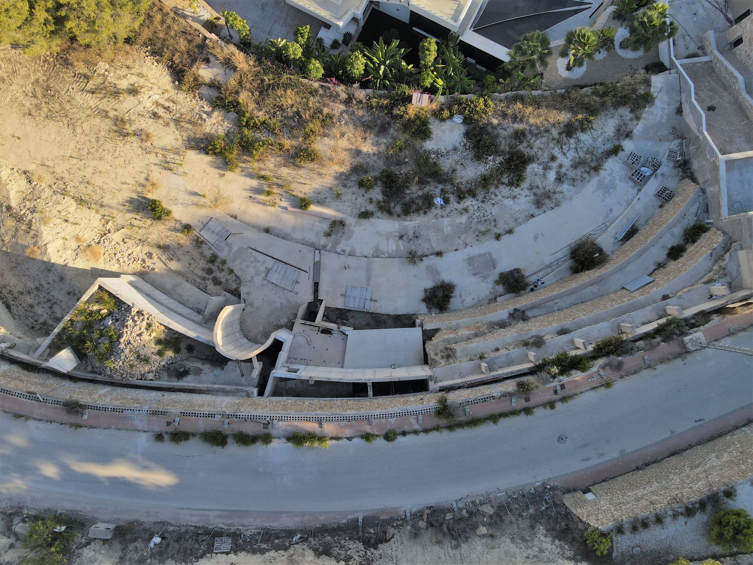 Luftaufnahme eines terrassierten Baugrundstücks mit geschwungenen Betonfundamenten und in den Hang integrierten Stützmauern aus Stein.