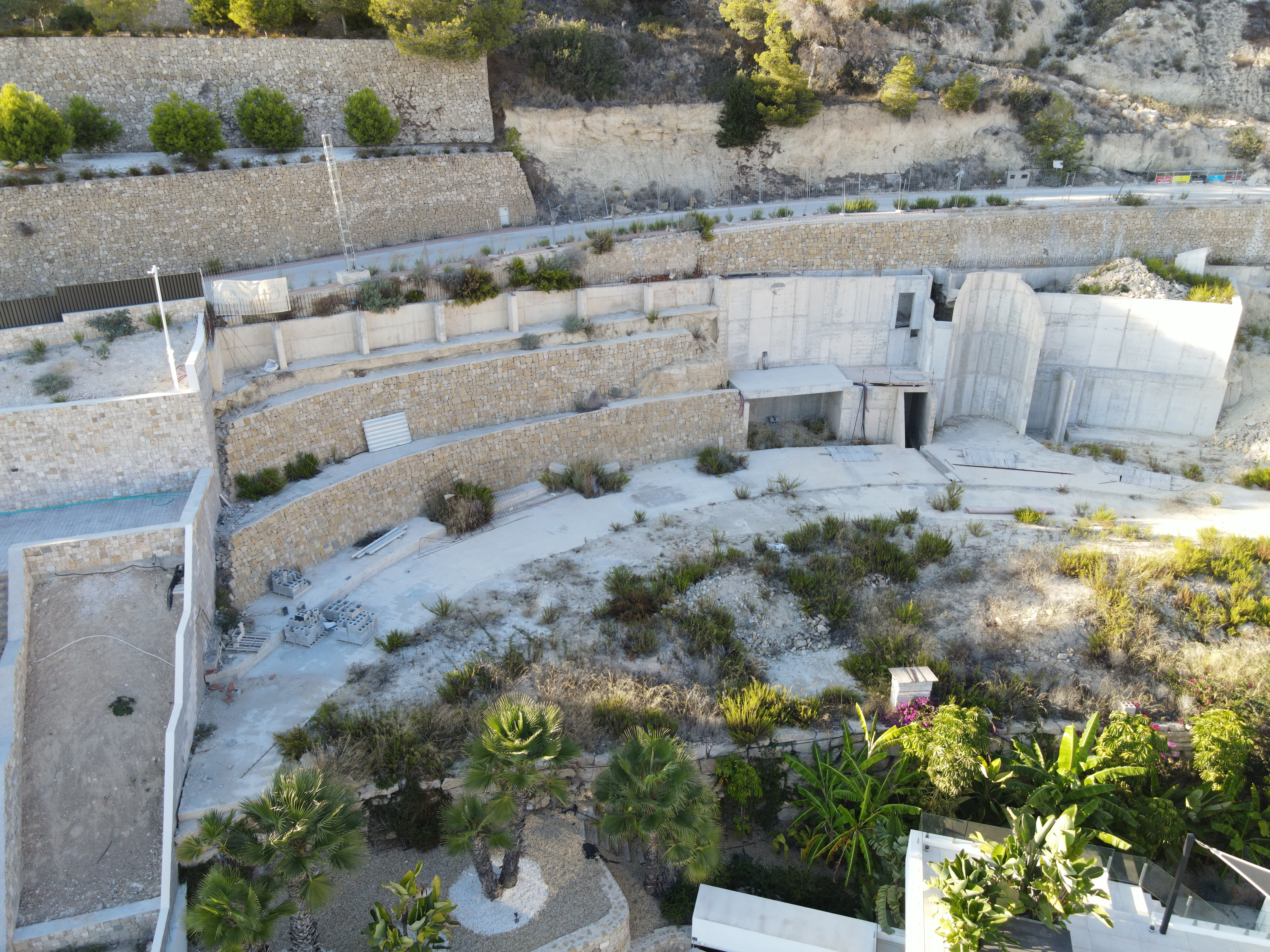 Luftaufnahme von terrassierten Steinmauern und Betonfundamenten, die in einen Hang mit Palmen und mediterraner Vegetation integriert sind.